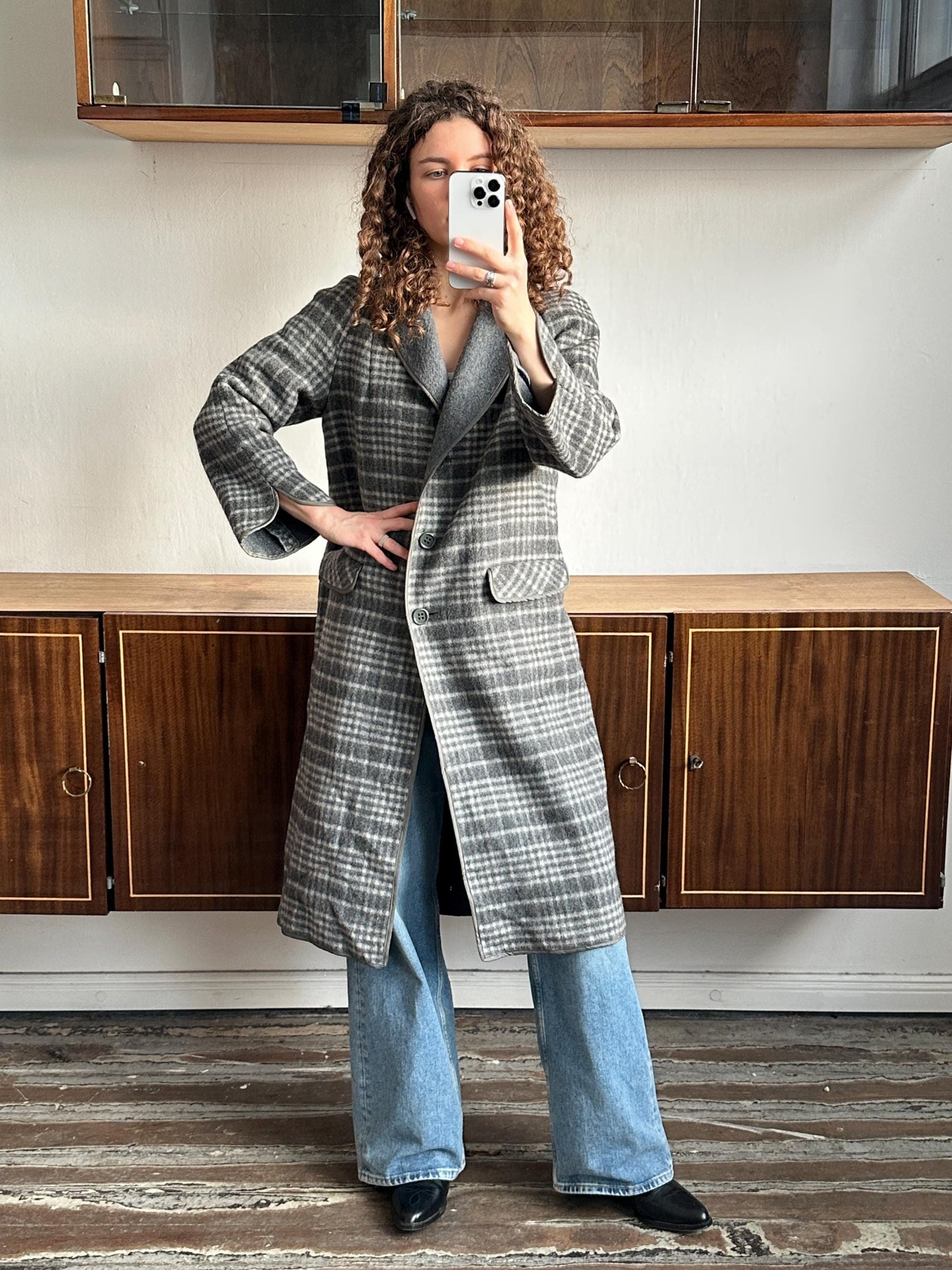 A woman with curly hair is taking a selfie with her phone, wearing a gray and white plaid coat and blue jeans.