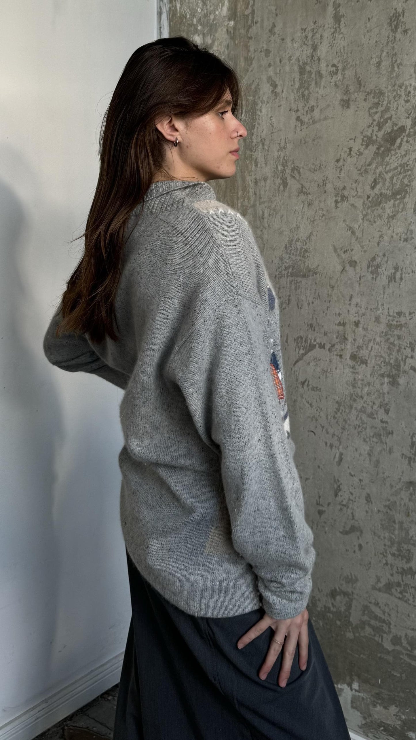 A woman with long brown hair stands in front of a concrete wall, wearing a gray sweater with a graphic design on the left side.