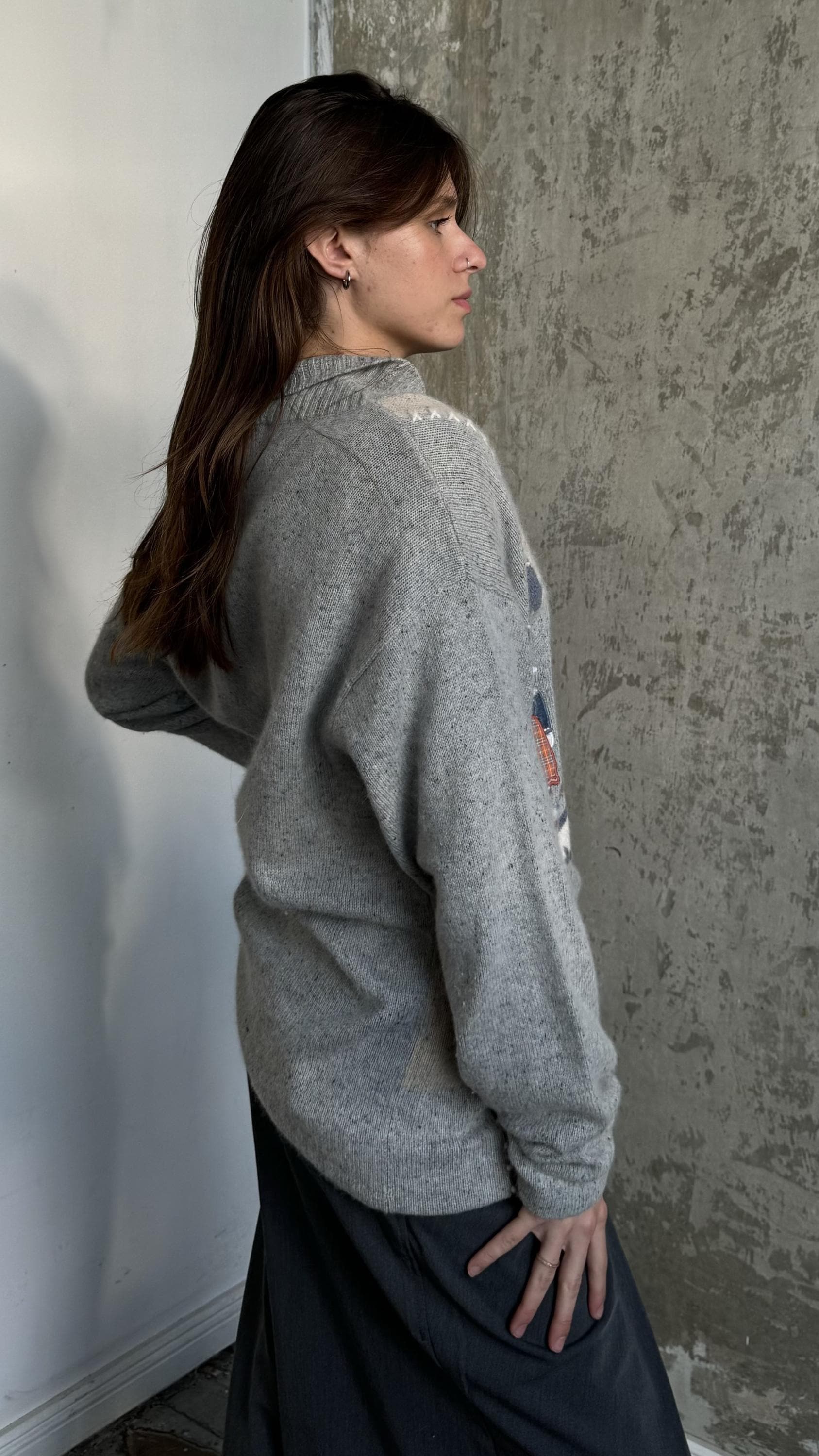 A woman with long brown hair stands in front of a concrete wall, wearing a gray sweater with a graphic design on the left side.