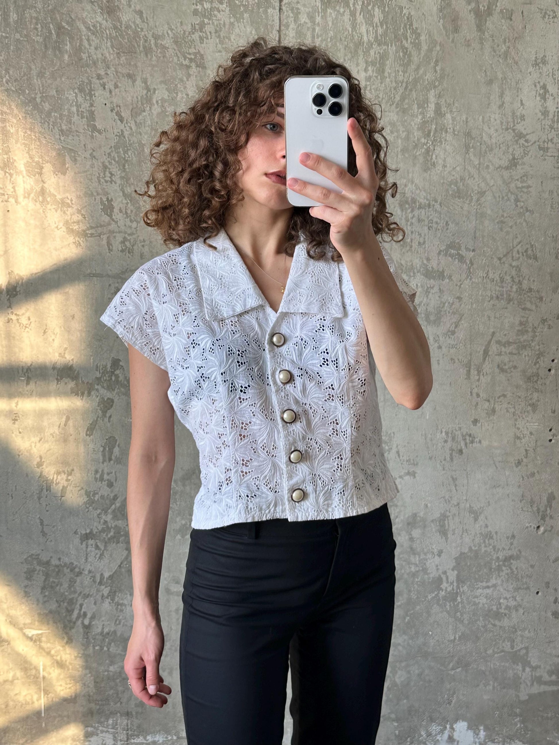 A woman with curly hair is taking a selfie with a white smartphone, wearing a white lace blouse with buttons and black pants.