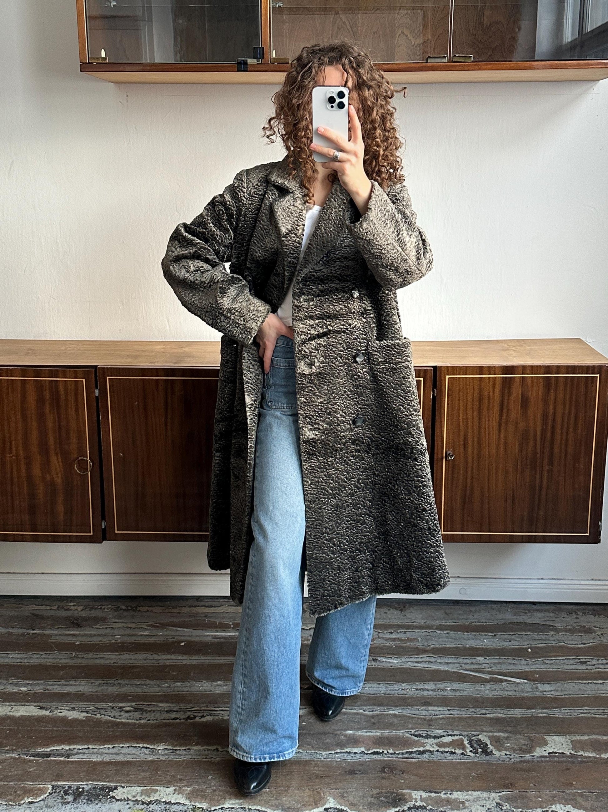 A person is taking a selfie with a smartphone, wearing a long coat and jeans, standing in front of a wooden cabinet.