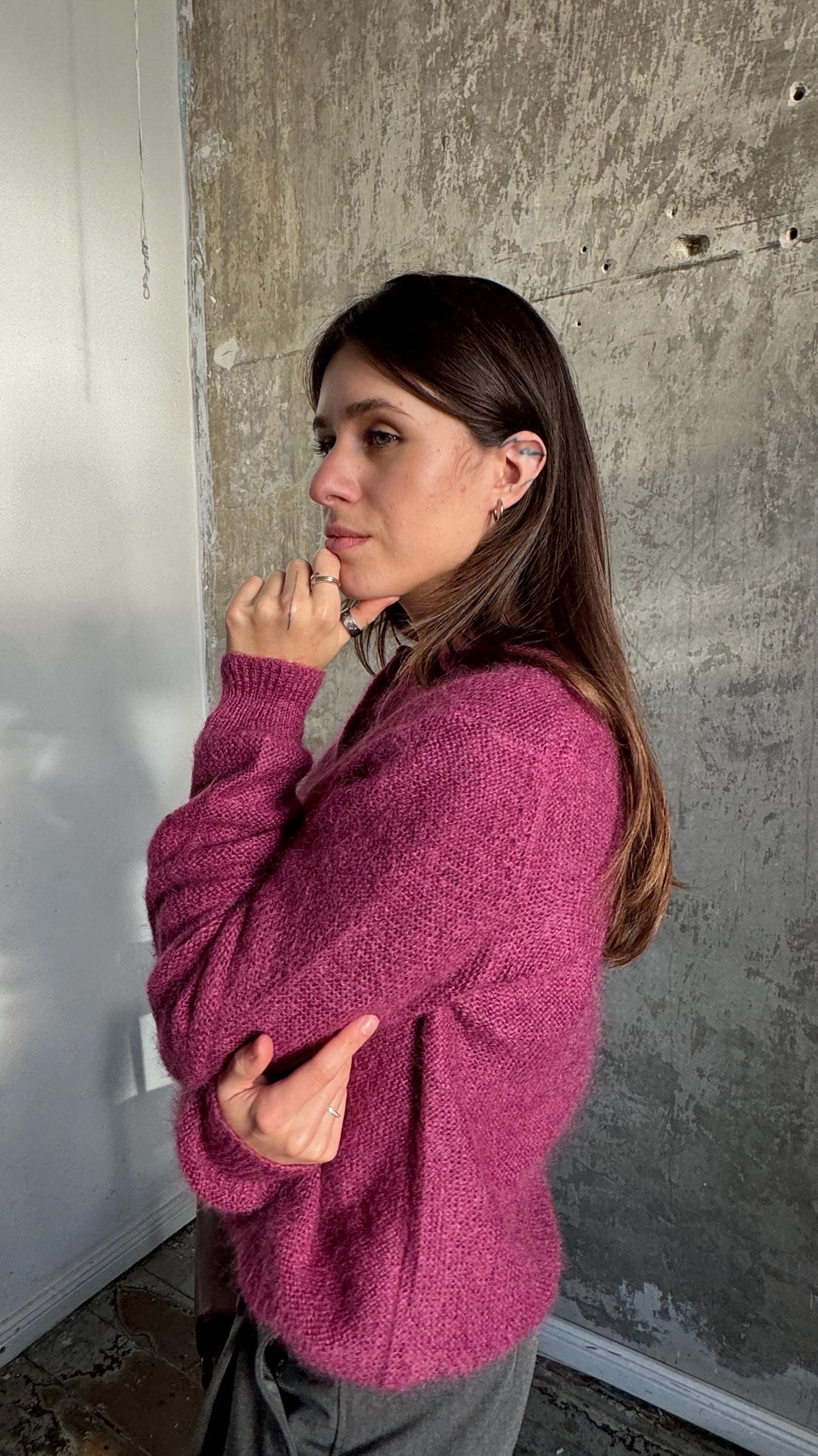 A woman wearing a pink sweater stands in front of a concrete wall, her hand resting on her chin as she gazes off to the side.