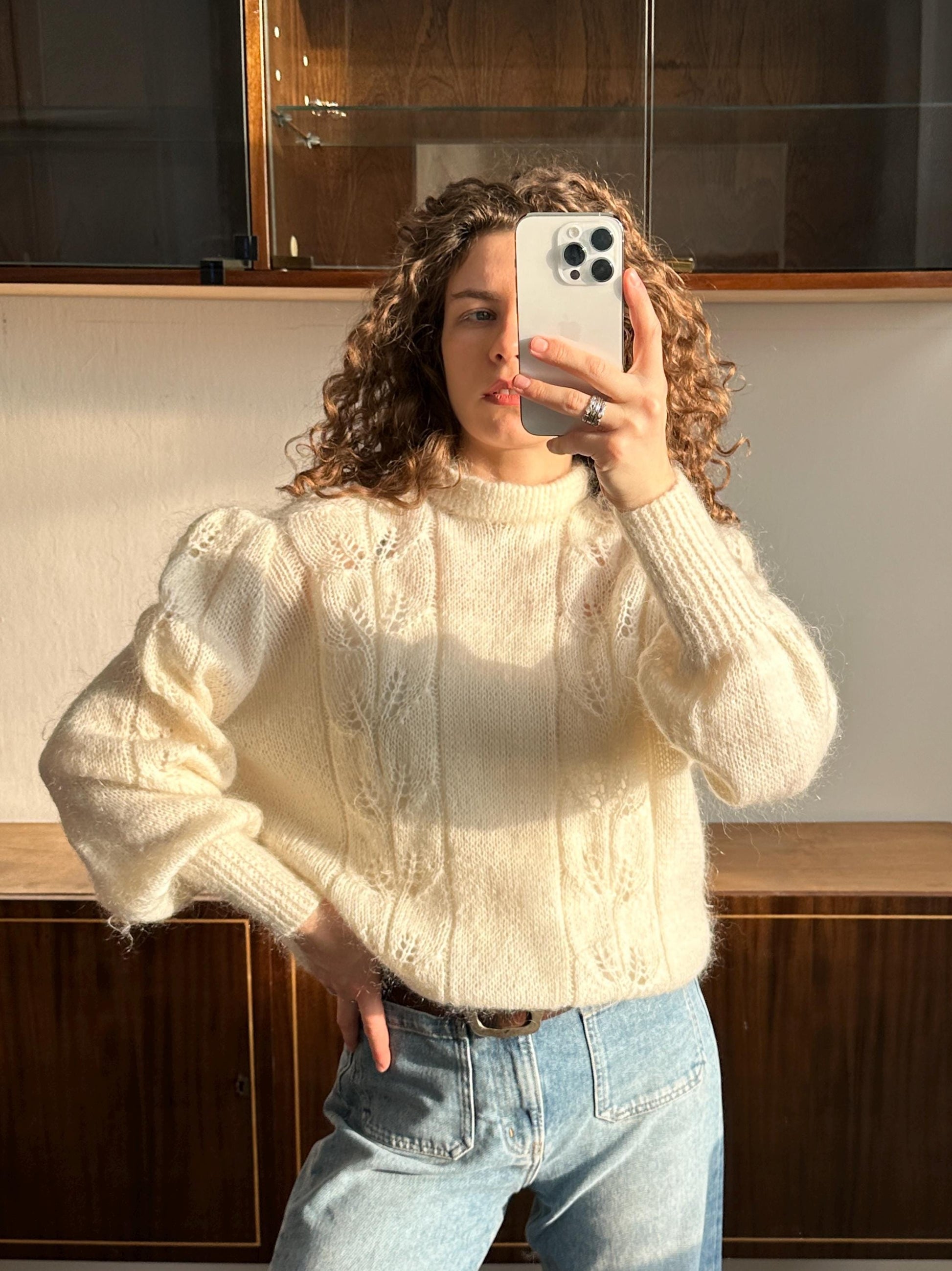 A woman with curly hair is taking a selfie with a white iPhone, wearing a white sweater and blue jeans.