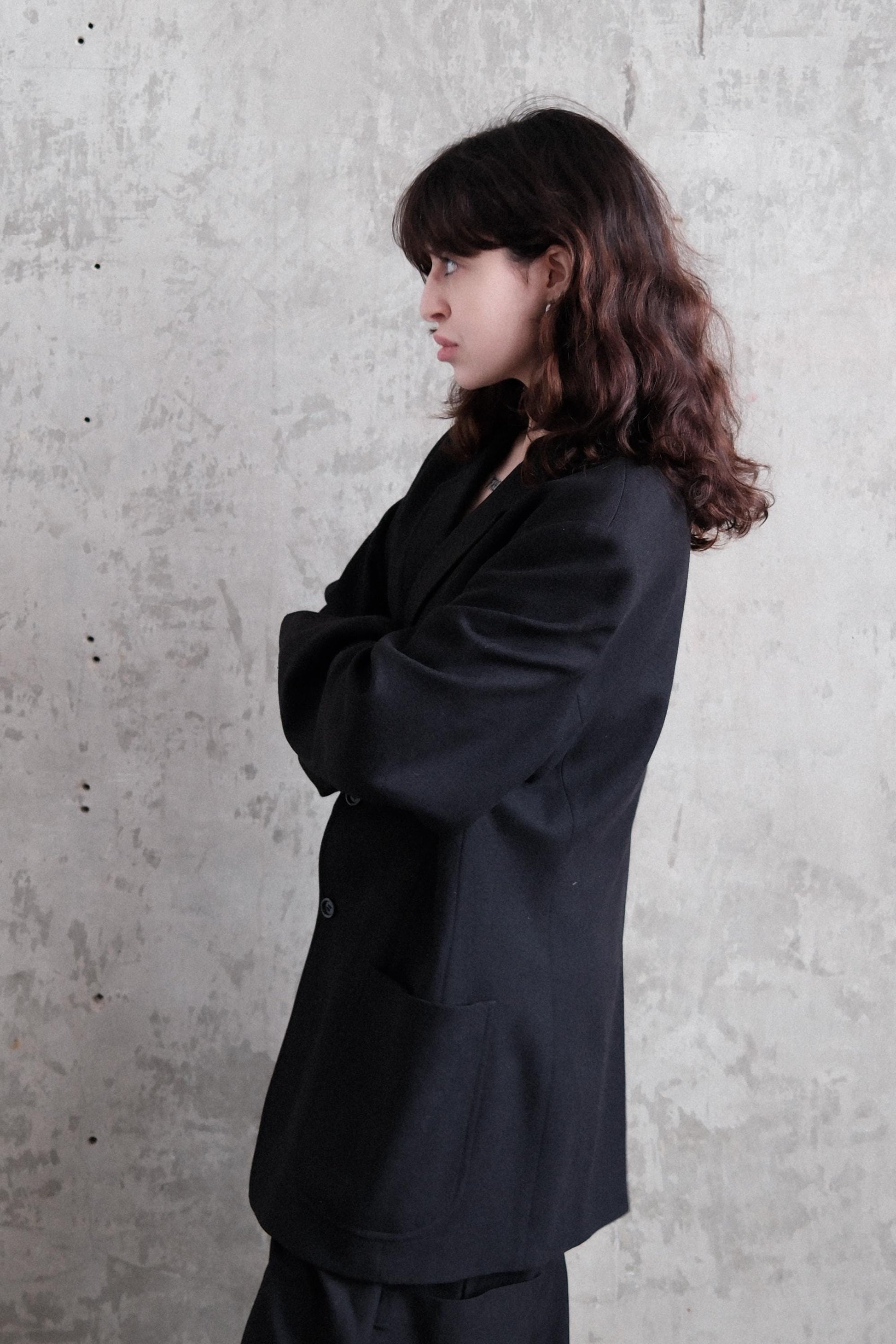 A woman with long brown hair is lying on her stomach, wearing a black jacket, against a plain concrete wall.