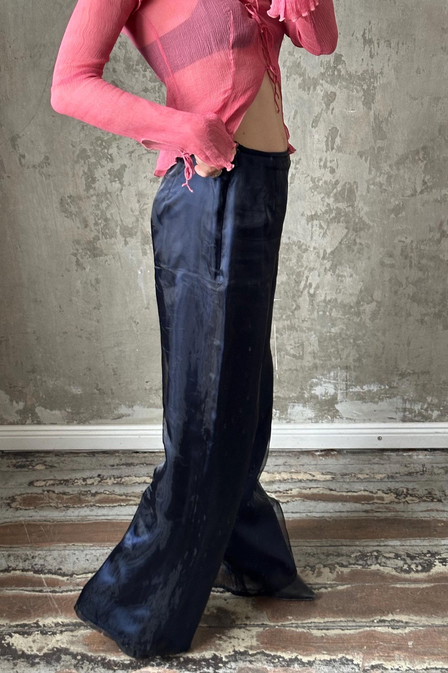 A person wearing a pink top and dark blue pants stands in front of a concrete wall.
