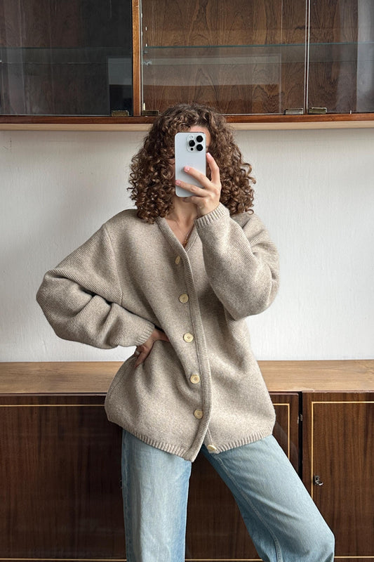 A person with curly hair is taking a selfie with a white phone, standing in front of a wooden cabinet.