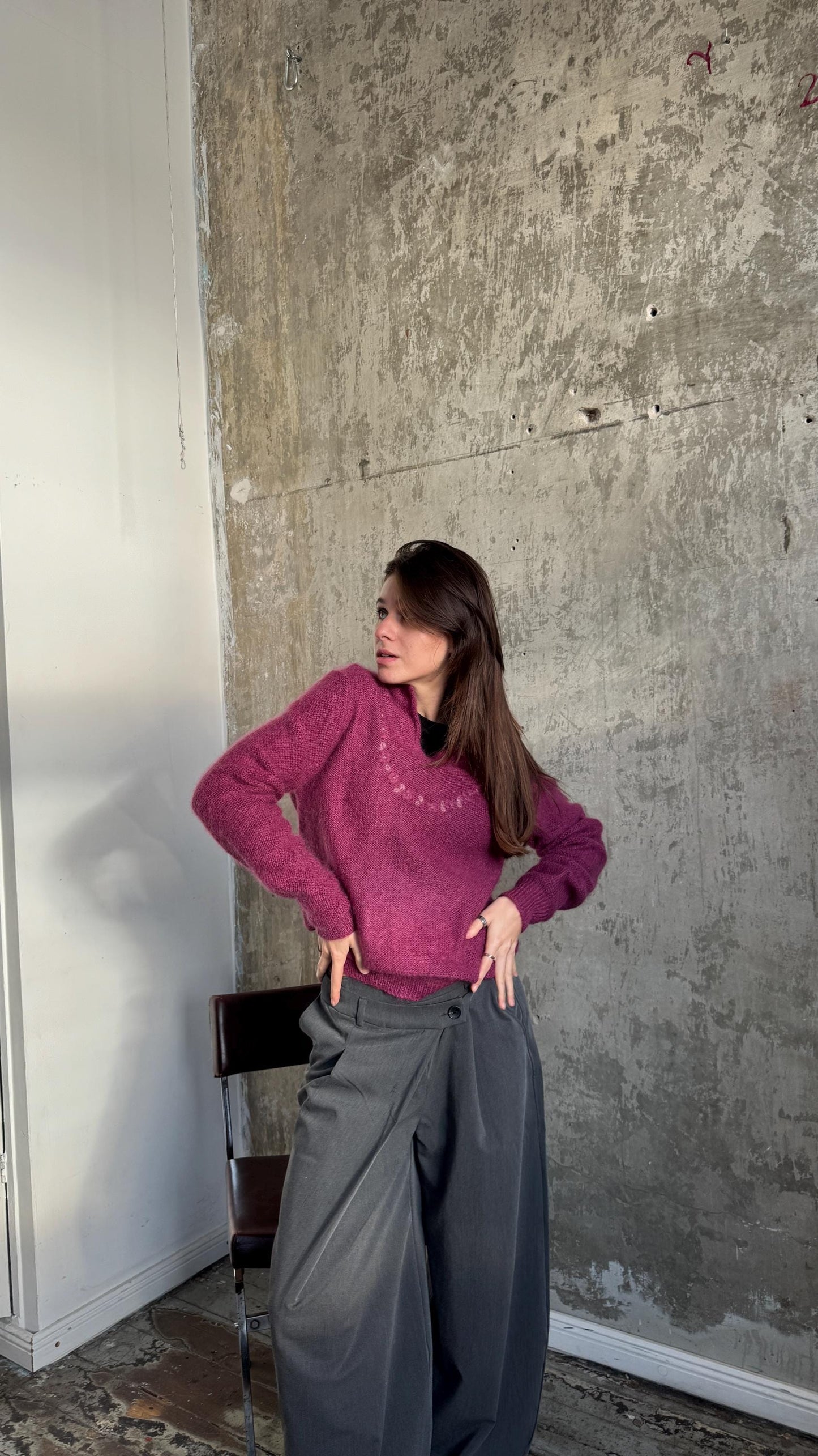 A woman wearing a pink sweater and gray pants stands in front of a concrete wall, with her hands on her hips.