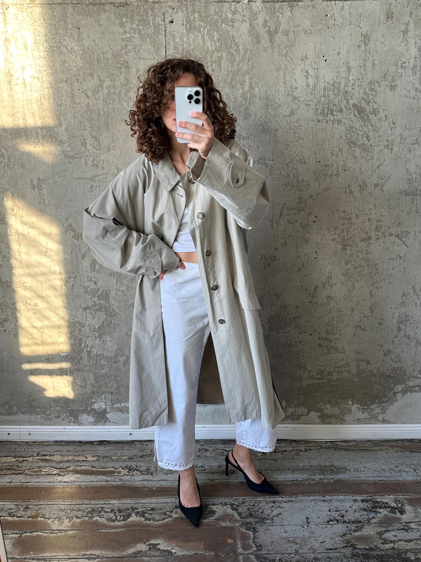 A woman in a beige trench coat and white pants is taking a selfie with a smartphone, standing in front of a concrete wall.