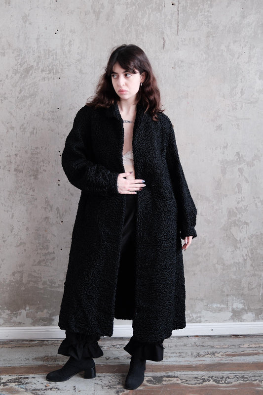 A woman wearing a long black coat stands against a plain gray wall.