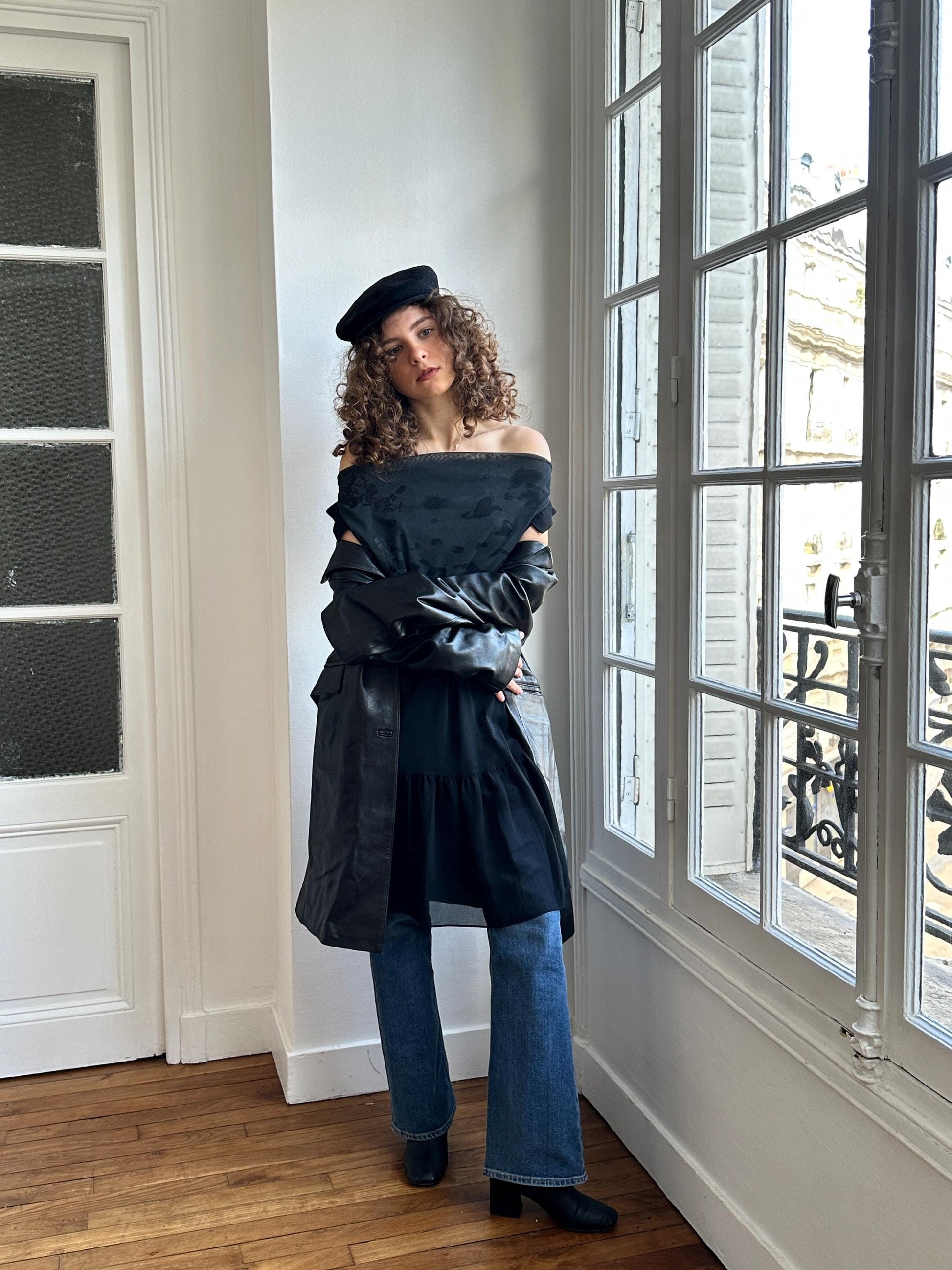A woman with curly hair stands in a room, wearing a black off-the-shoulder top, a black leather jacket, and blue jeans. She is posing with her arms crossed and leaning against a window.