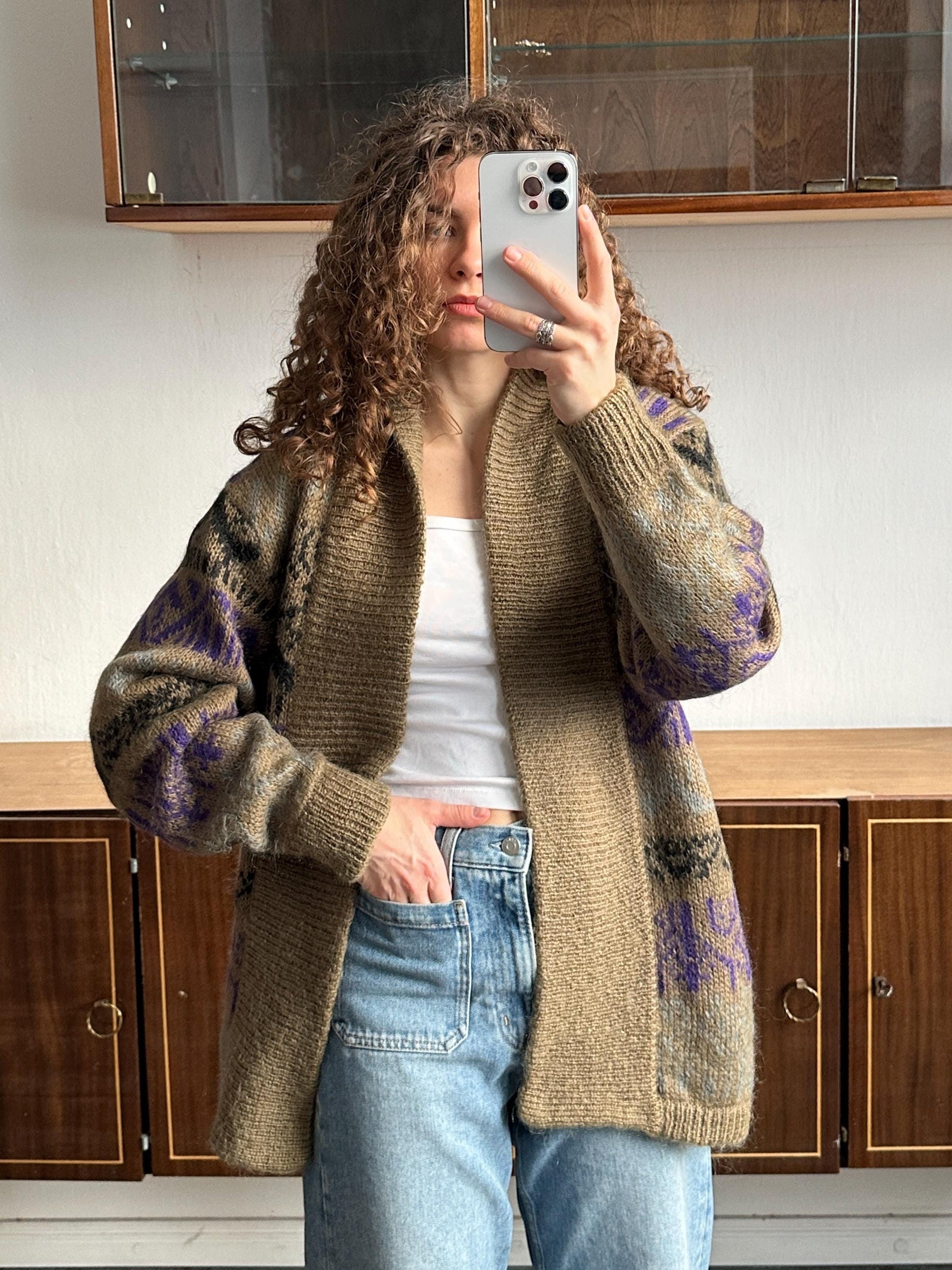 A woman with curly hair is taking a selfie with her iPhone, wearing a brown and purple patterned cardigan and blue jeans.