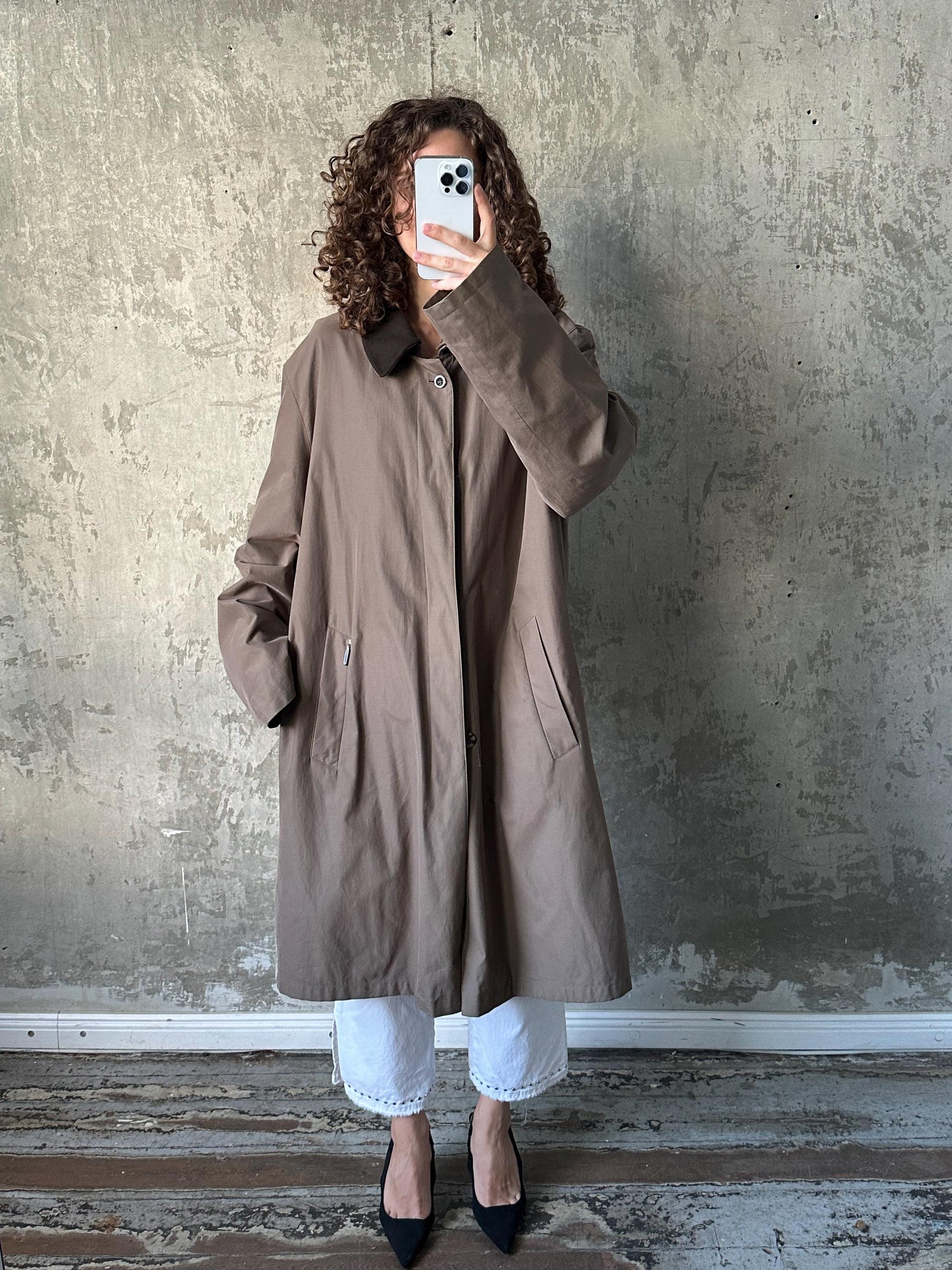 A person with curly hair is taking a selfie with a smartphone, wearing a brown coat and white pants, standing in front of a concrete wall.