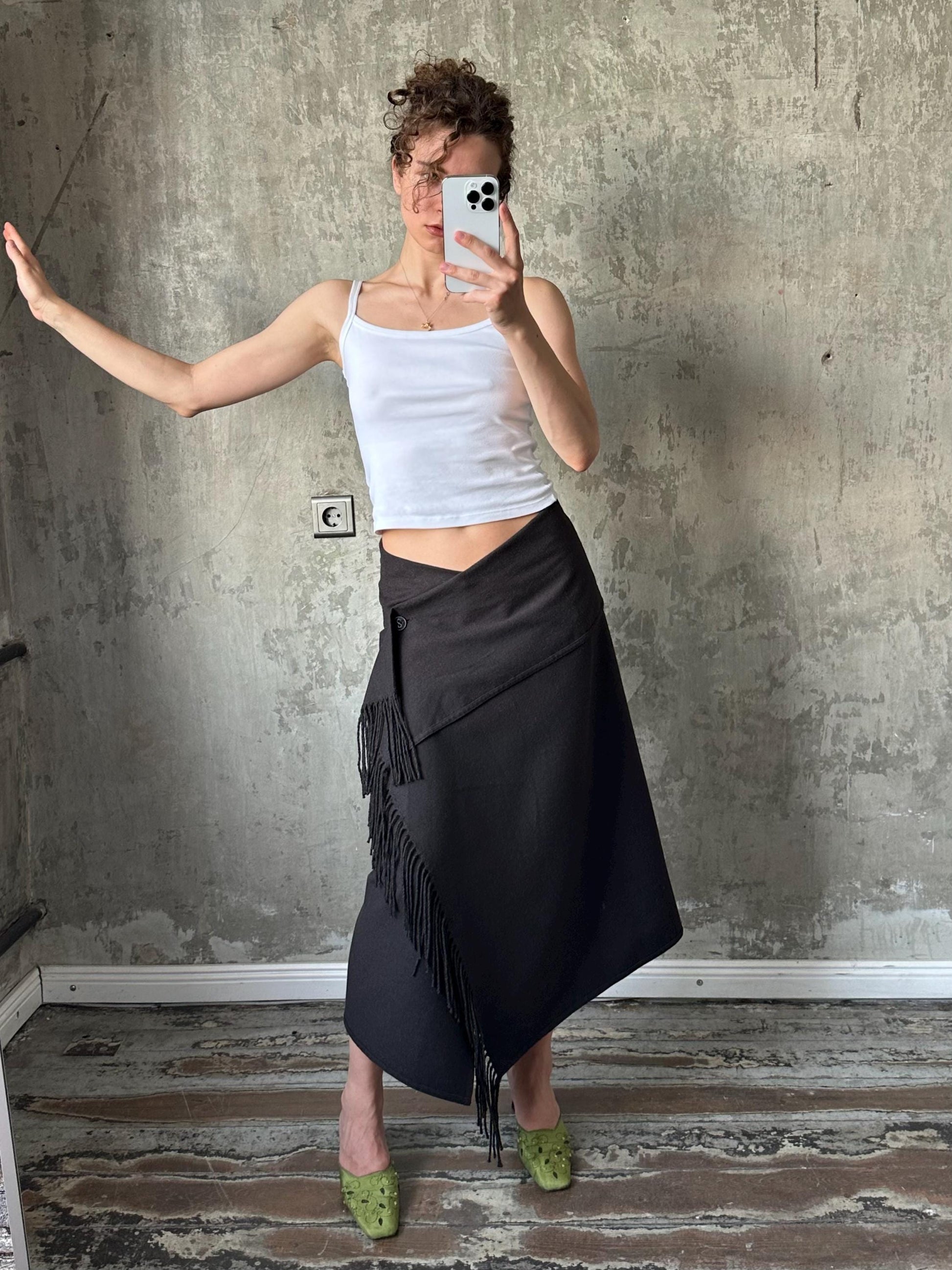 A woman is taking a selfie with her phone, wearing a white tank top and a black skirt with fringe details.