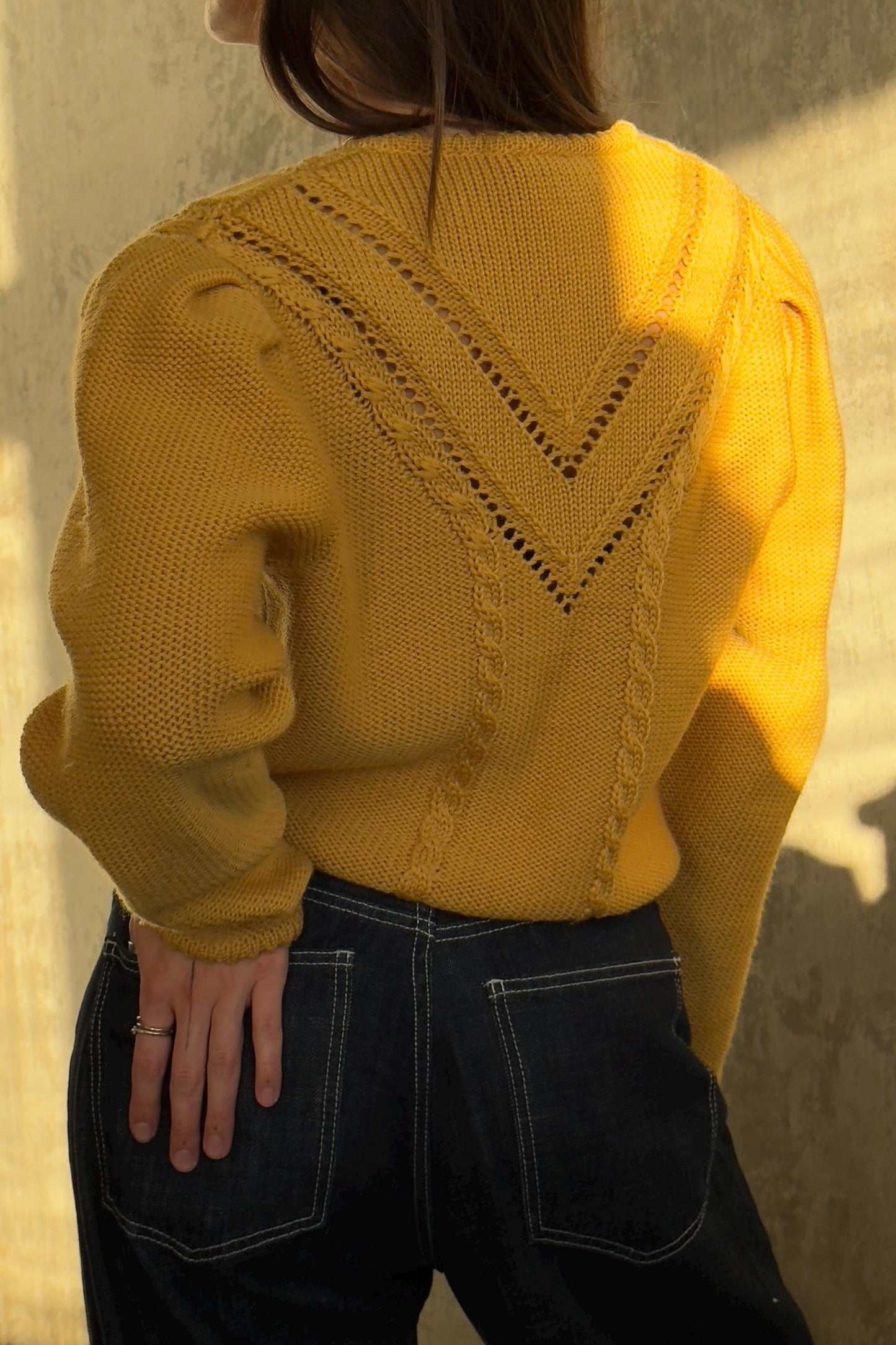 A person wearing a yellow sweater with a geometric pattern and dark jeans stands against a concrete wall.