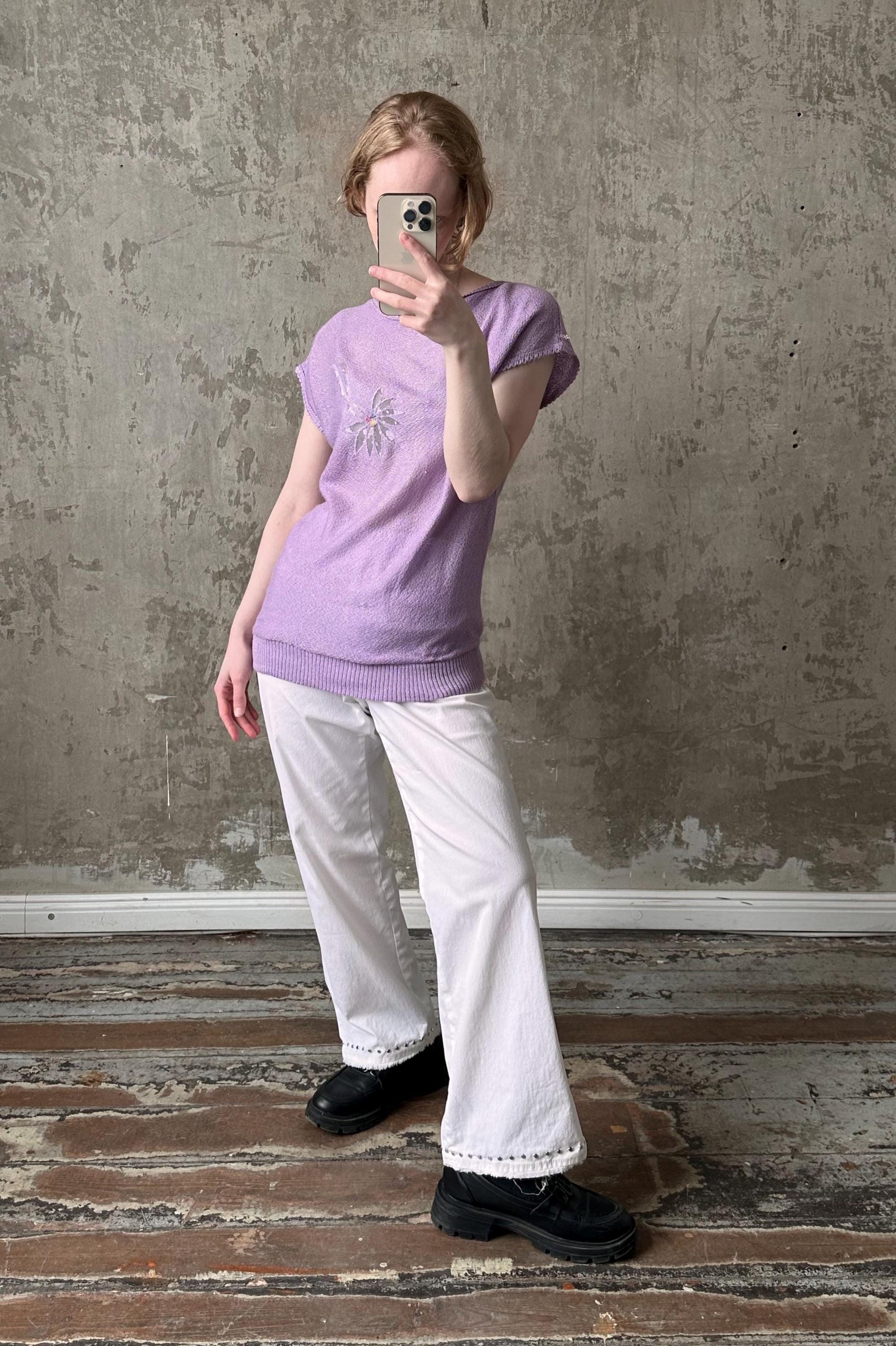 A person is taking a selfie with a smartphone, wearing a purple top and white pants, standing in front of a concrete wall.