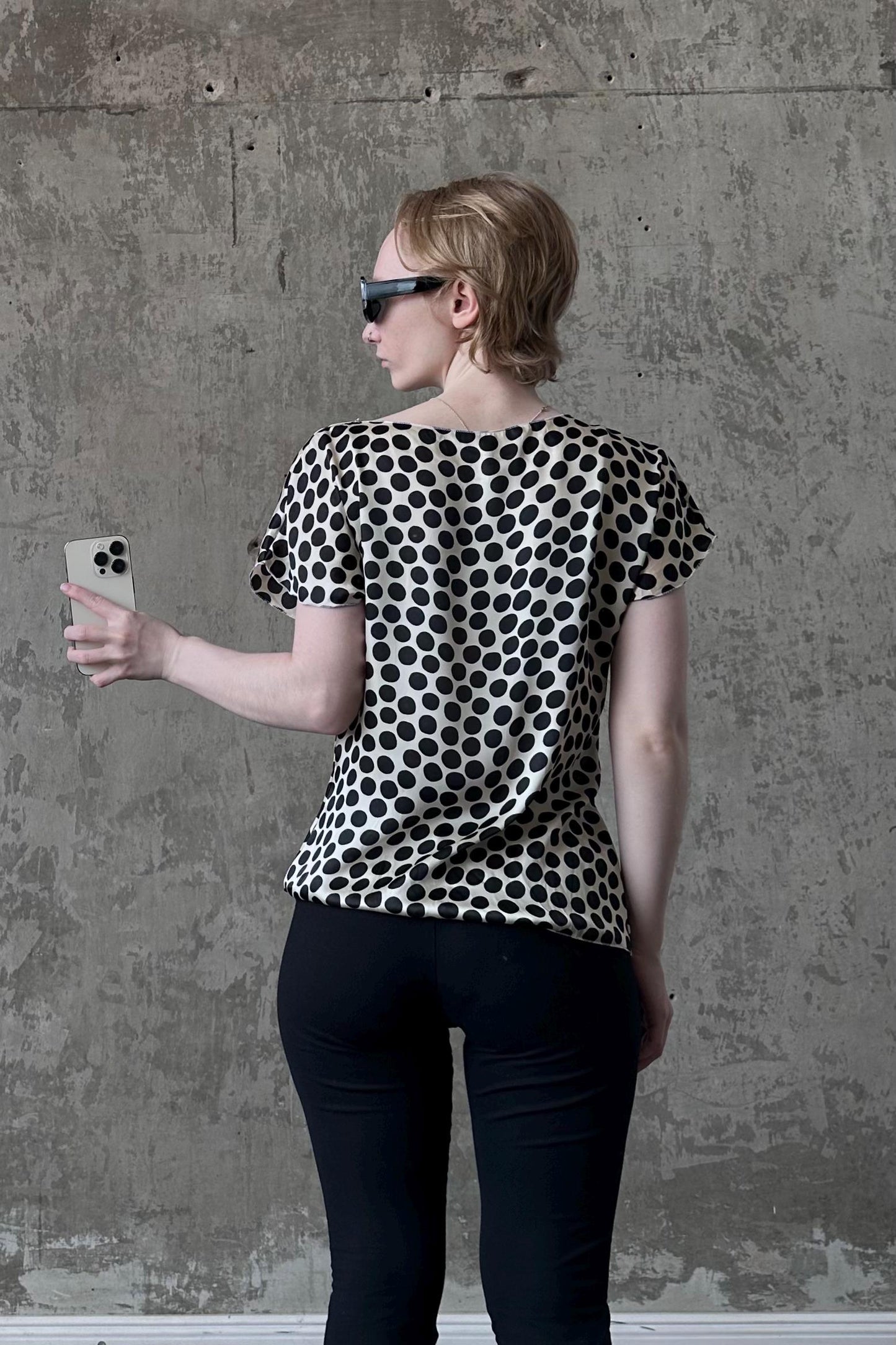 A woman wearing a black and white polka dot blouse and sunglasses is standing in front of a concrete wall, holding a smartphone.
