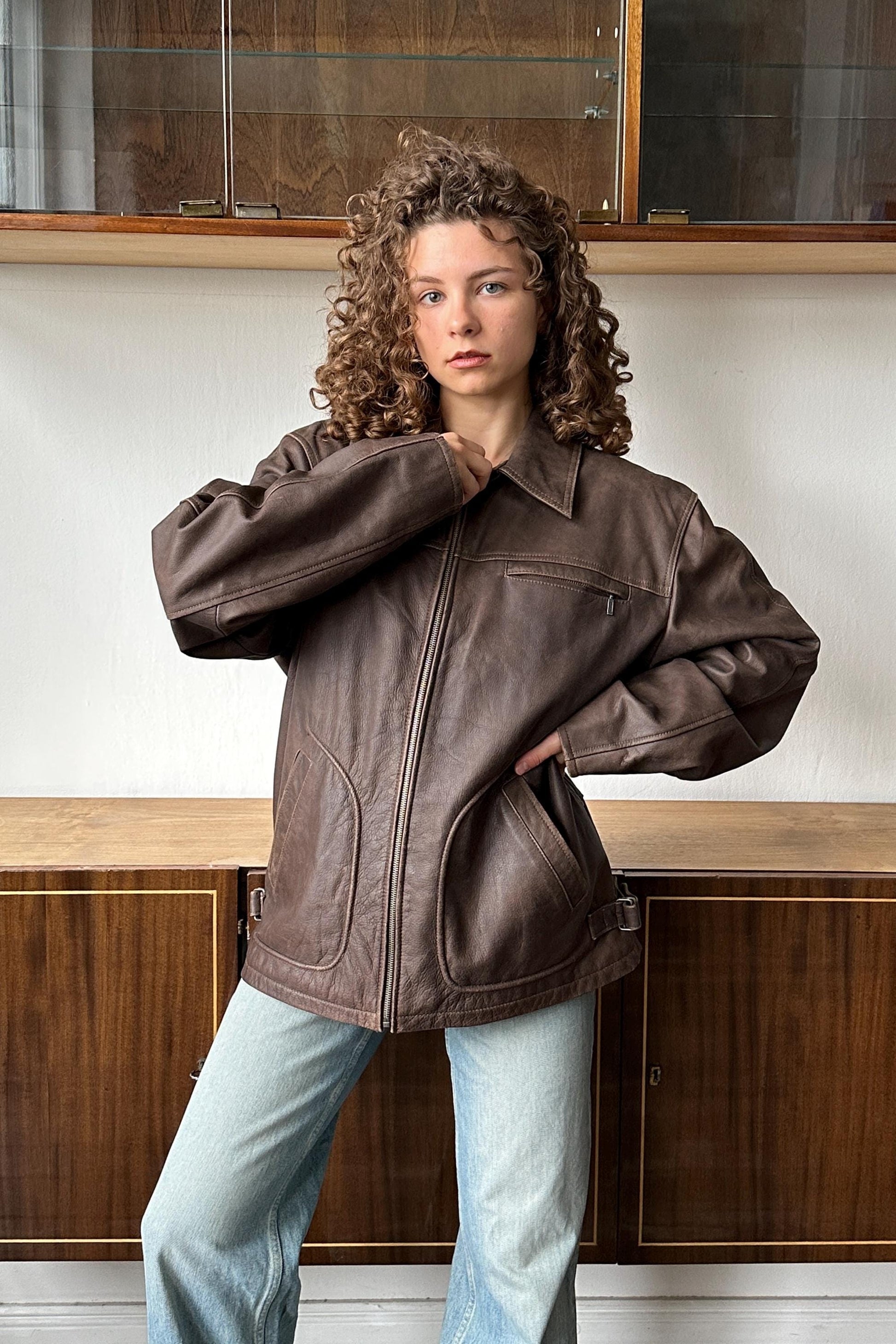 A young woman with curly hair wearing a brown leather jacket stands in front of a wooden cabinet.