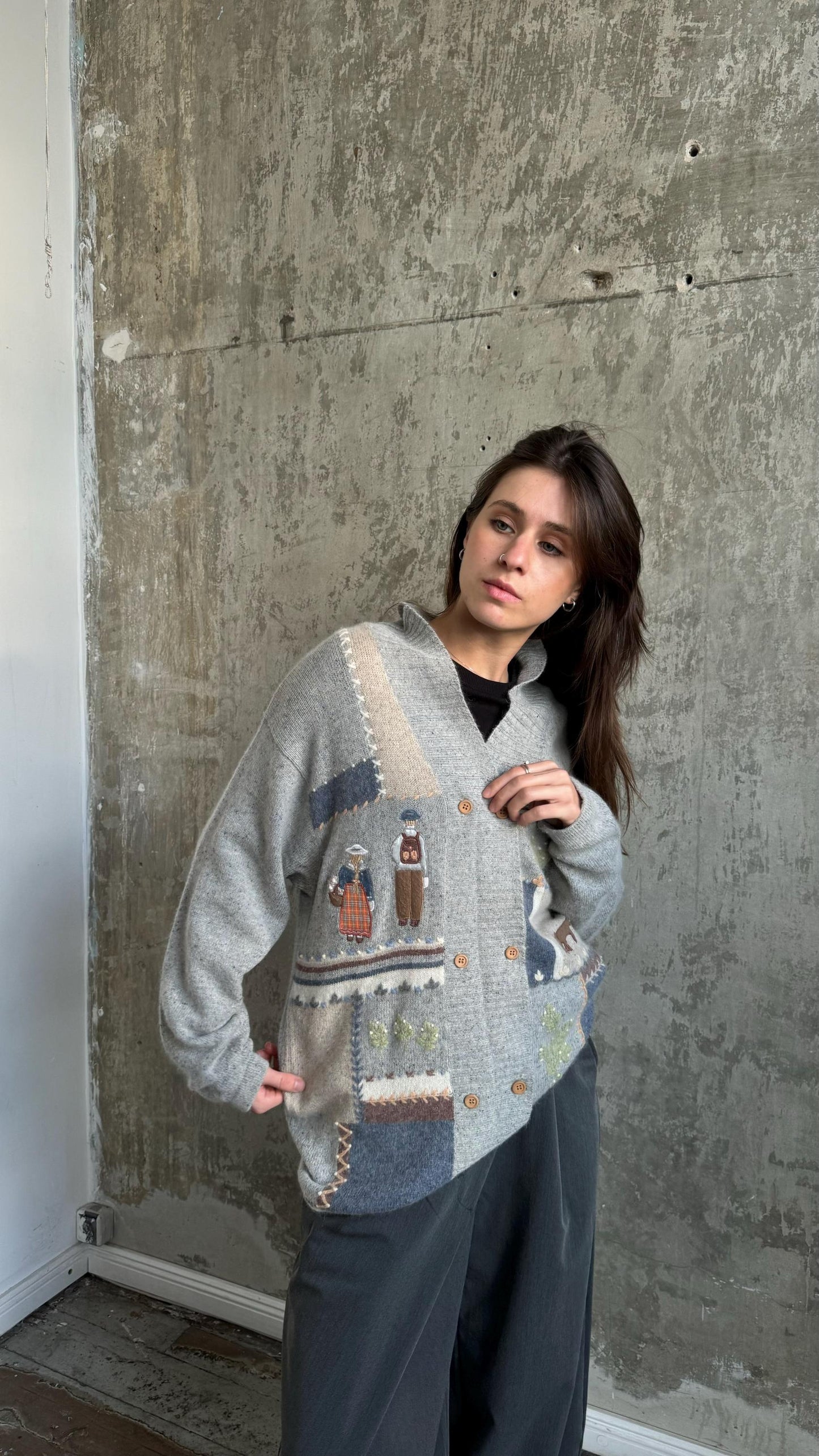 A woman wearing a gray cardigan with a patchwork design stands in front of a concrete wall.