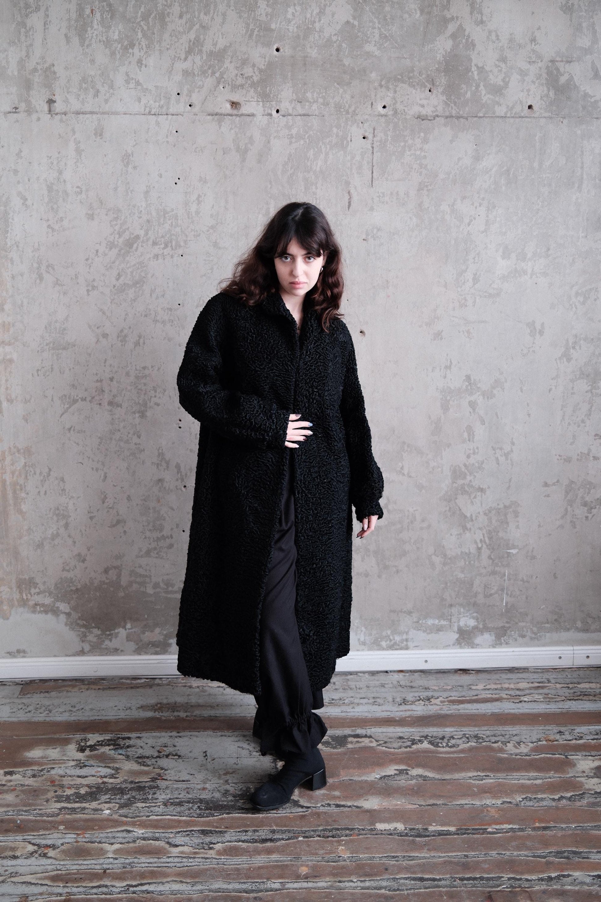 A woman in a long black coat stands in a room with a concrete wall and wooden floor.