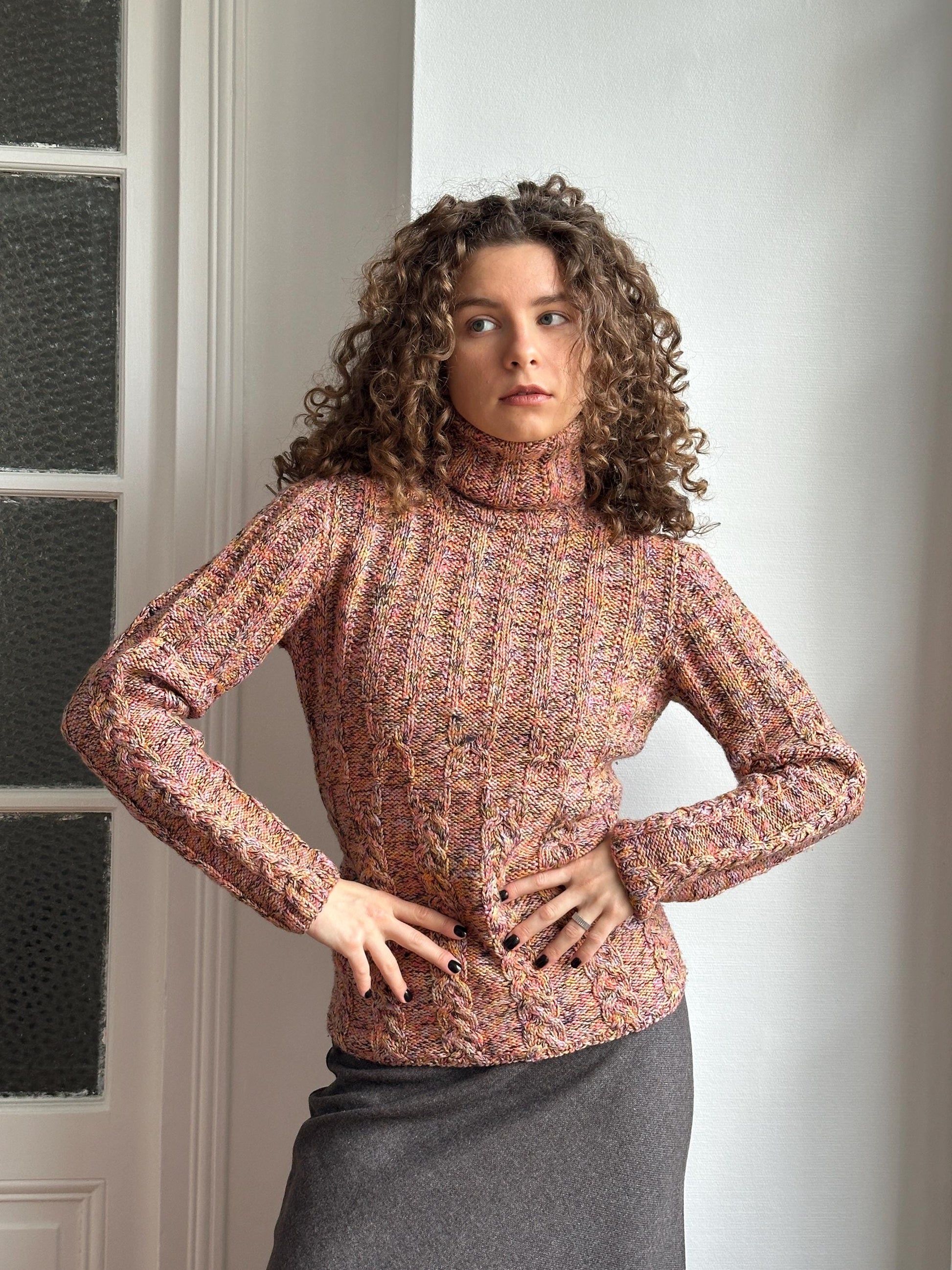 A woman with curly hair wearing a pink and white patterned sweater is posing with her hands on her hips, looking off to the side.