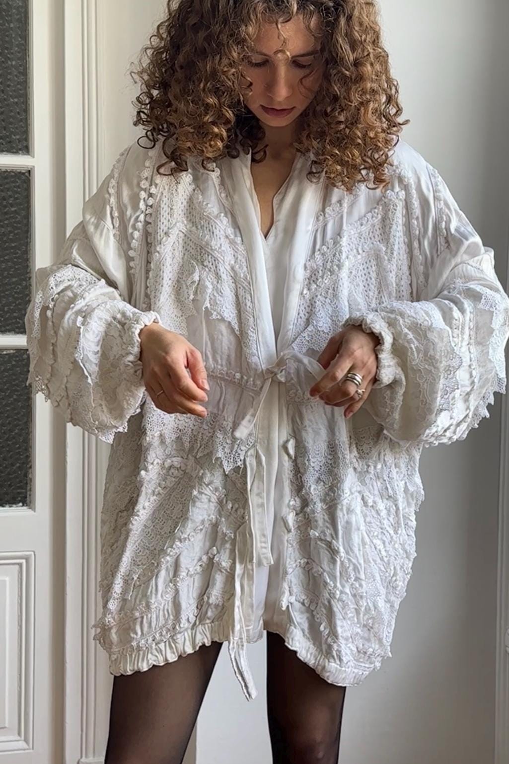 A woman with curly hair wearing a white, lace-trimmed robe stands in front of a white door.