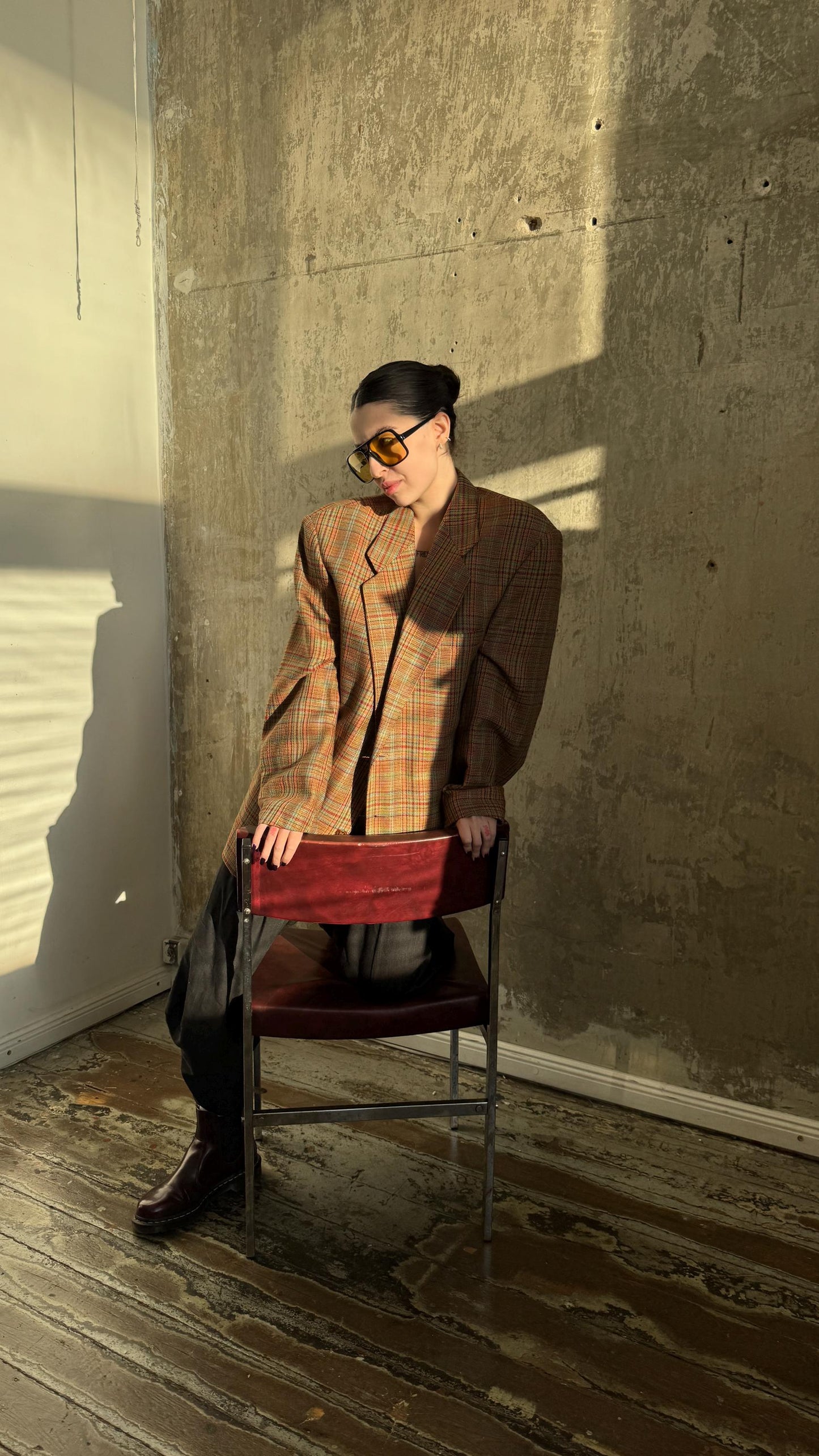 A person wearing a plaid jacket and sunglasses is sitting on a red chair in a room with a concrete wall.