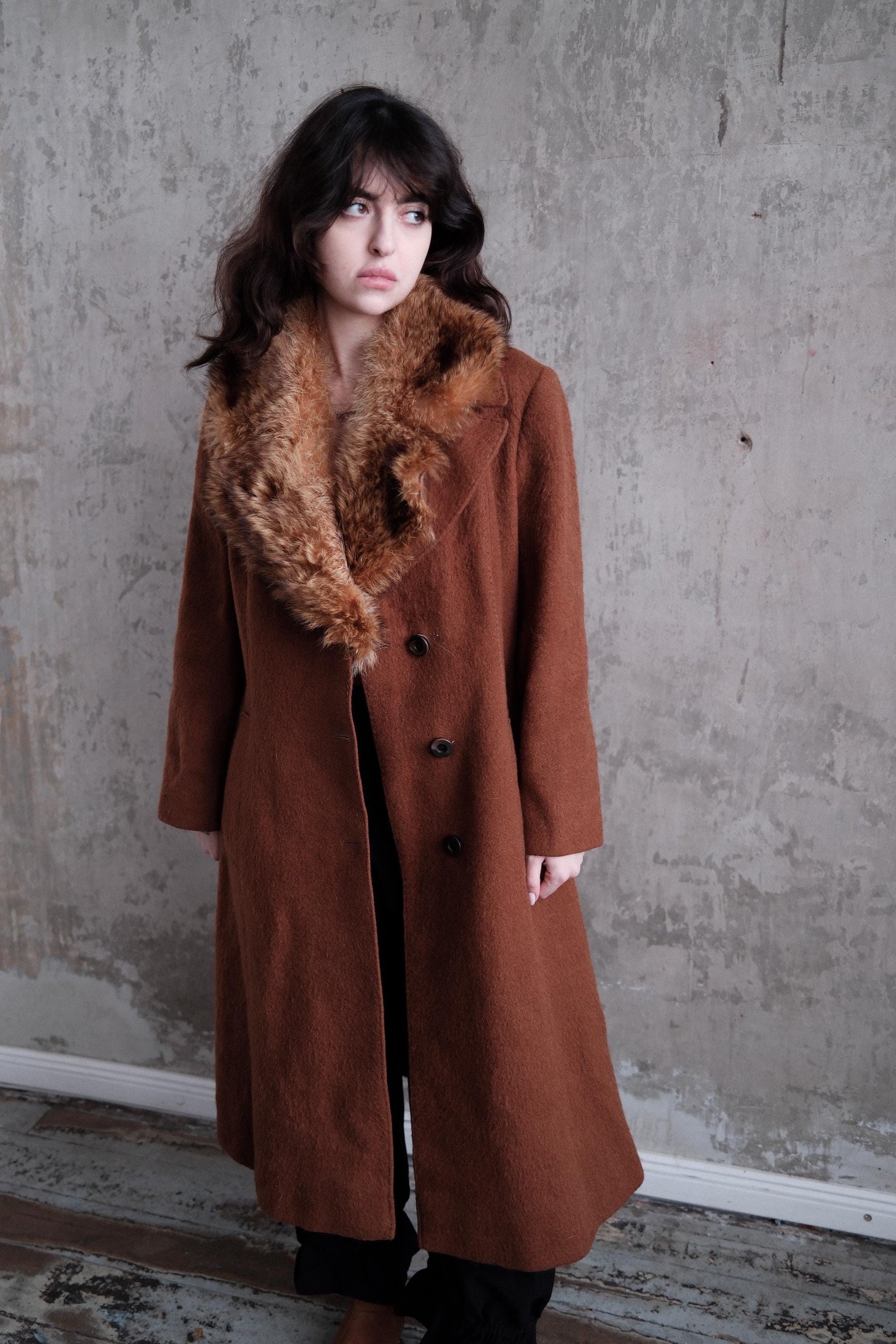A woman wearing a brown coat with a fur collar stands against a concrete wall.