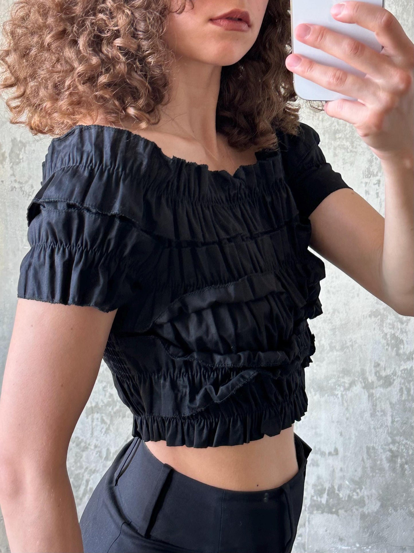 A woman with curly hair is taking a selfie with a smartphone, wearing a black ruffled off-the-shoulder top.