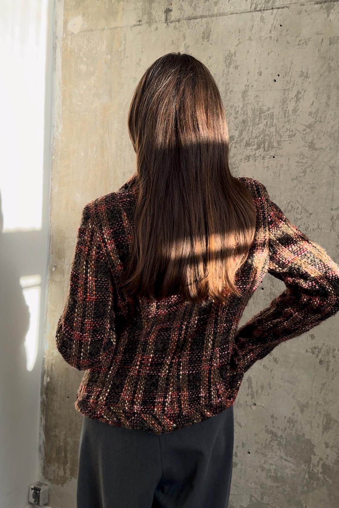 A person with long brown hair stands in front of a concrete wall, wearing a colorful, patterned sweater.