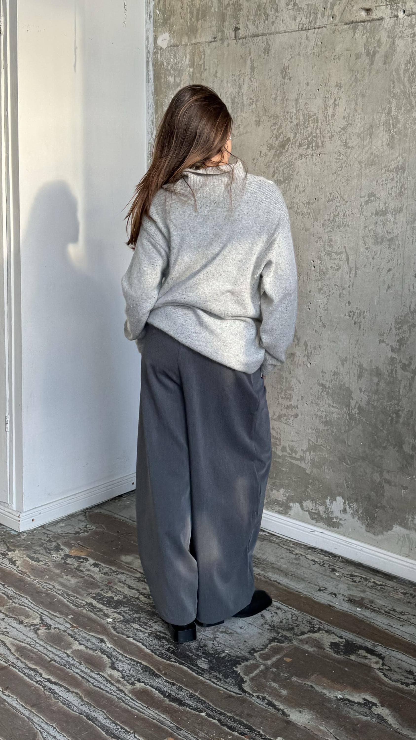 A person wearing a gray sweater and gray pants stands in a room with a concrete wall and wooden floor.