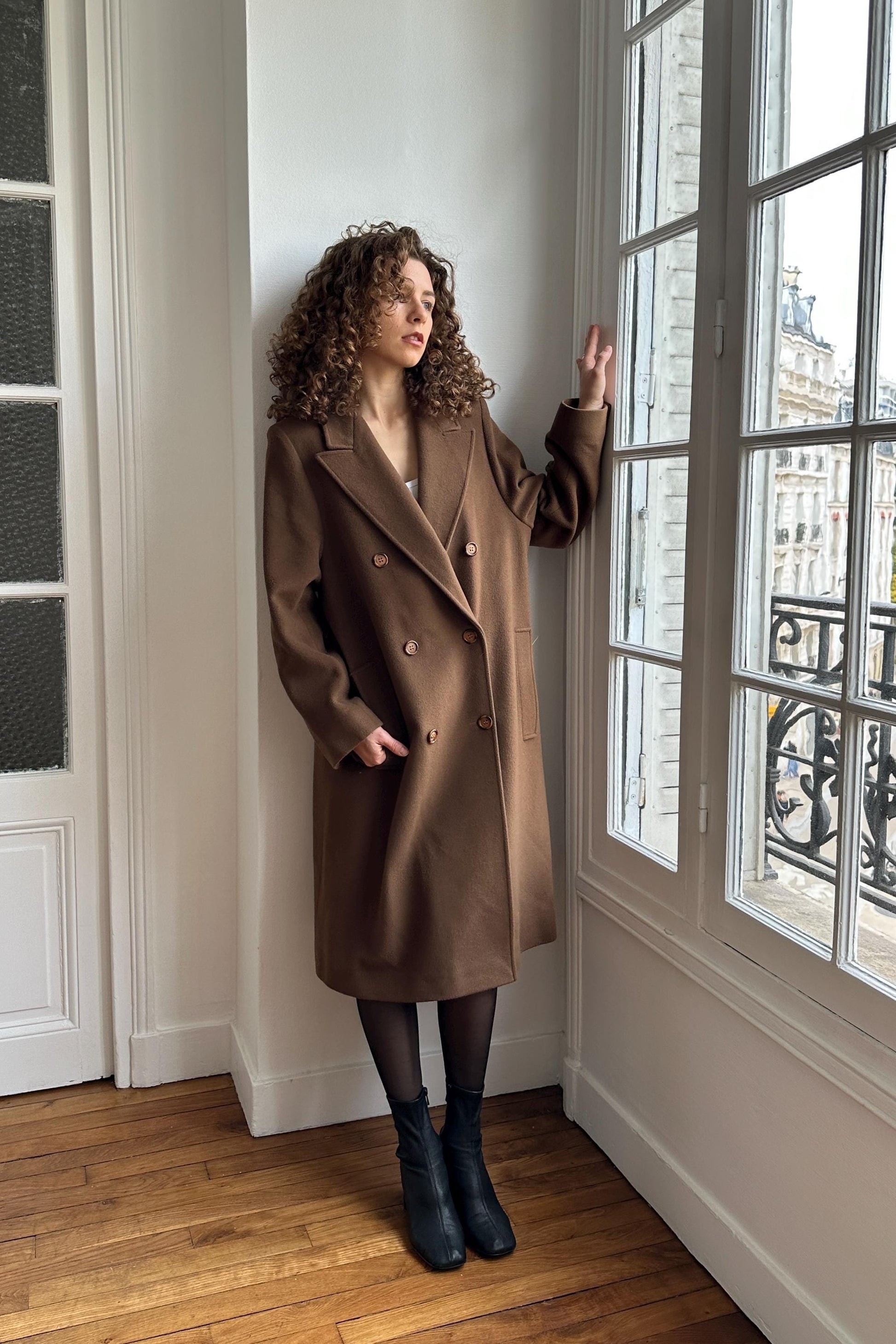 A woman in a brown coat stands in a room with a window, looking out at the cityscape.