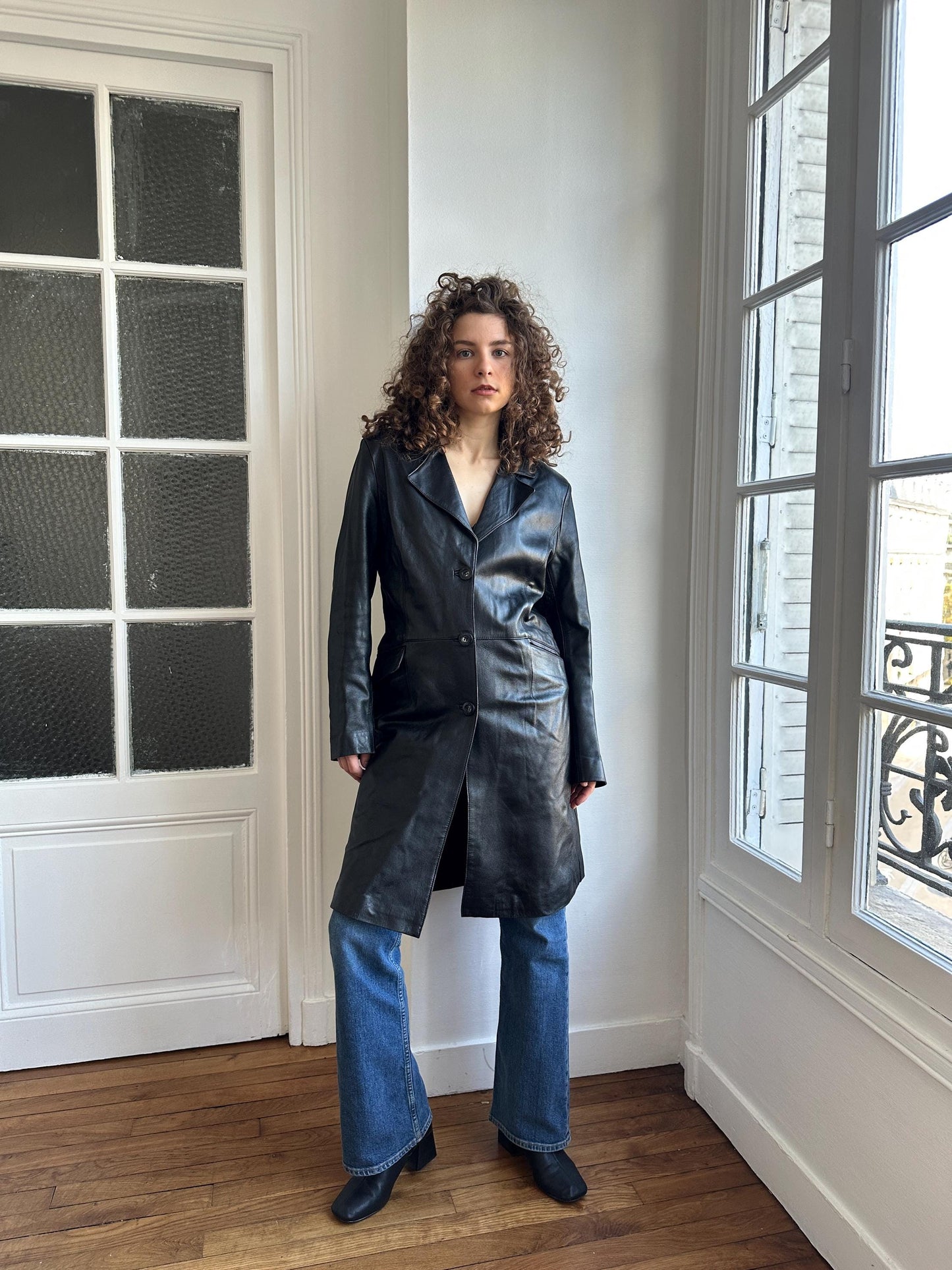 A woman wearing a black leather coat stands in a room with a window and a door.