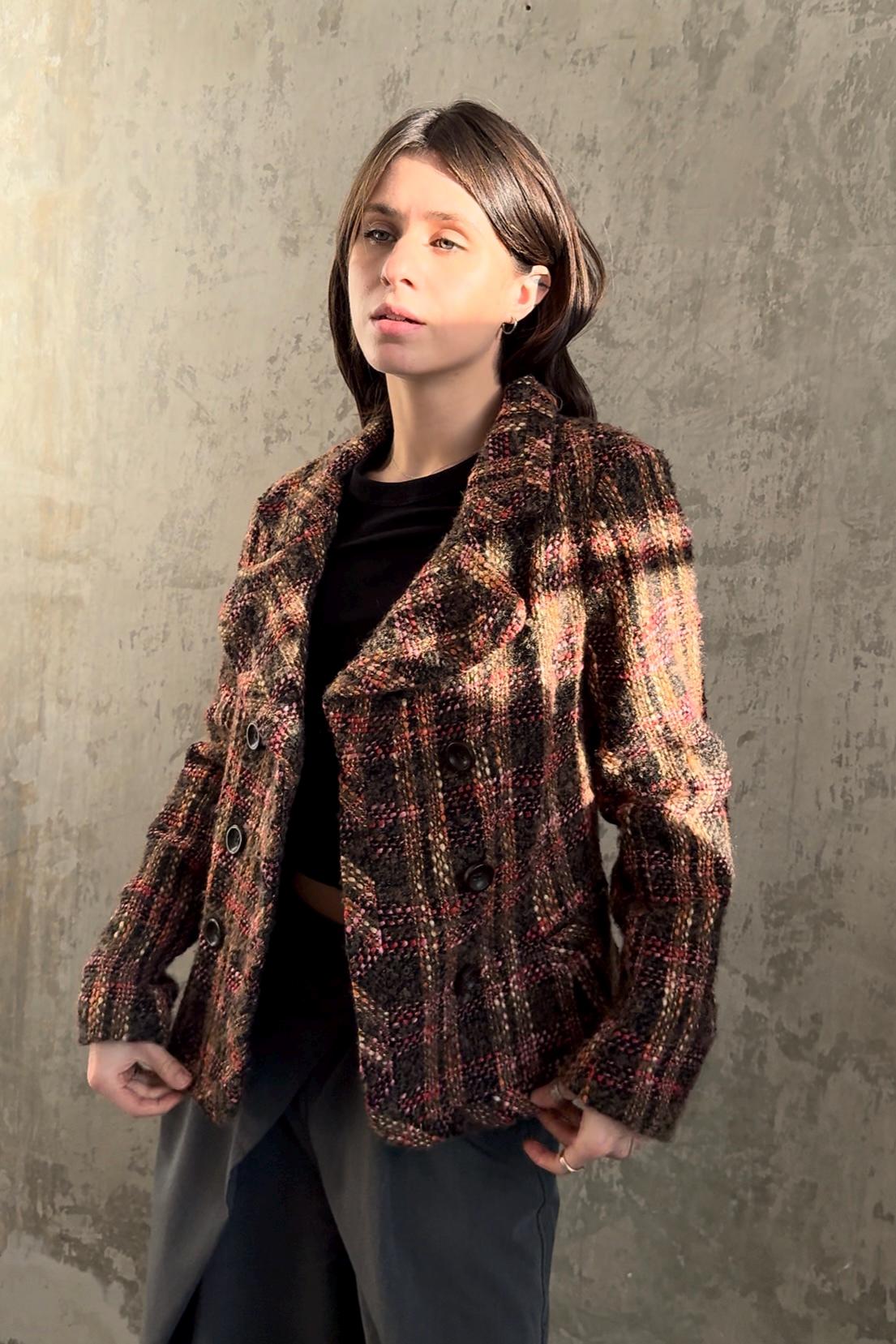 A young woman wearing a brown and black plaid jacket stands against a concrete wall, looking directly at the camera with a serious expression.