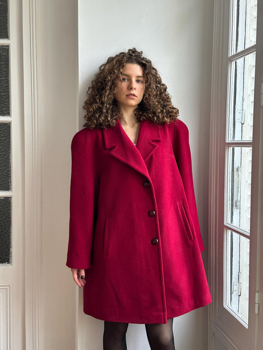 A person wearing a red coat is leaning against a wall, with their head tilted back and looking to the side.