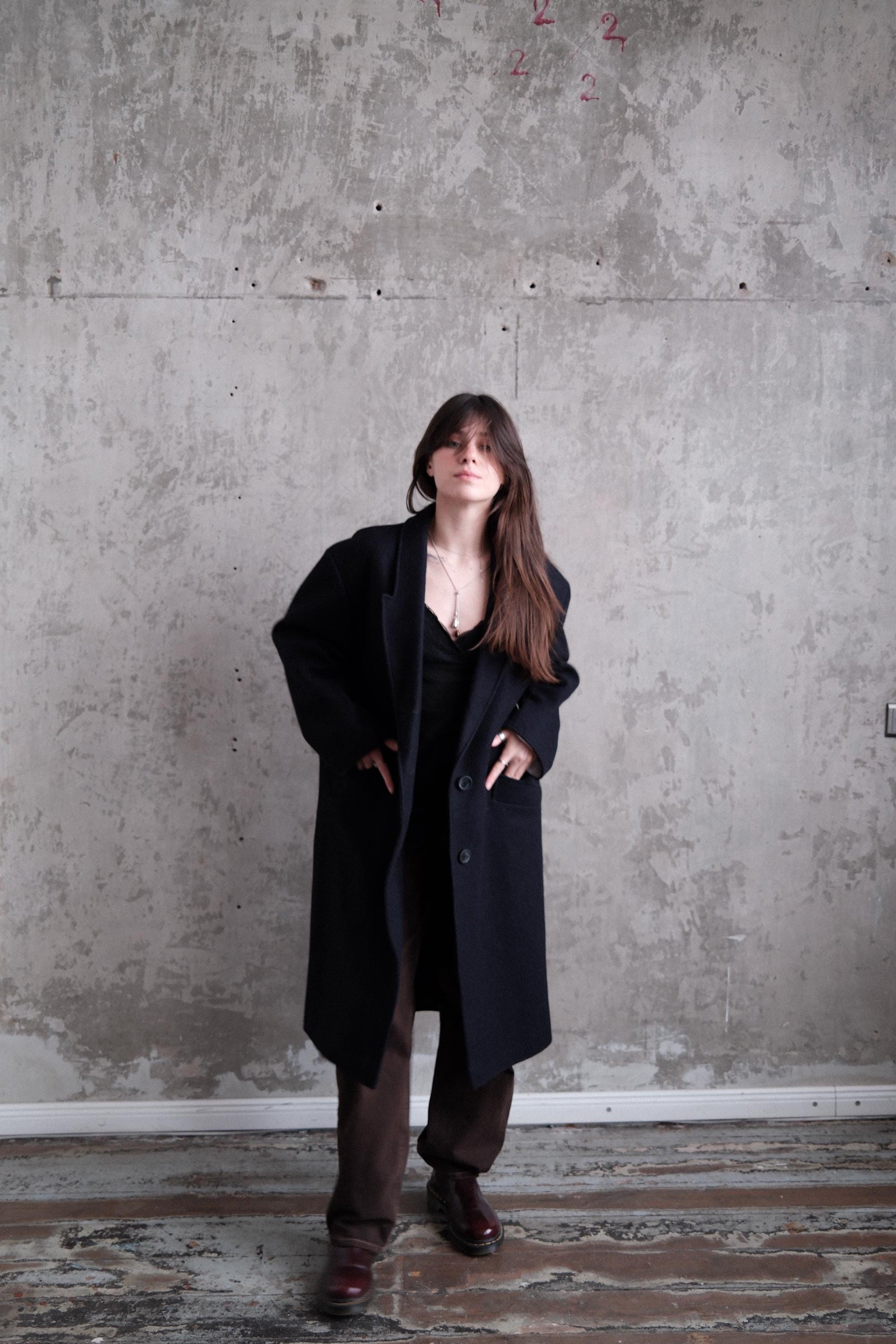 A person wearing a black coat is standing in a room with a concrete wall in the background.