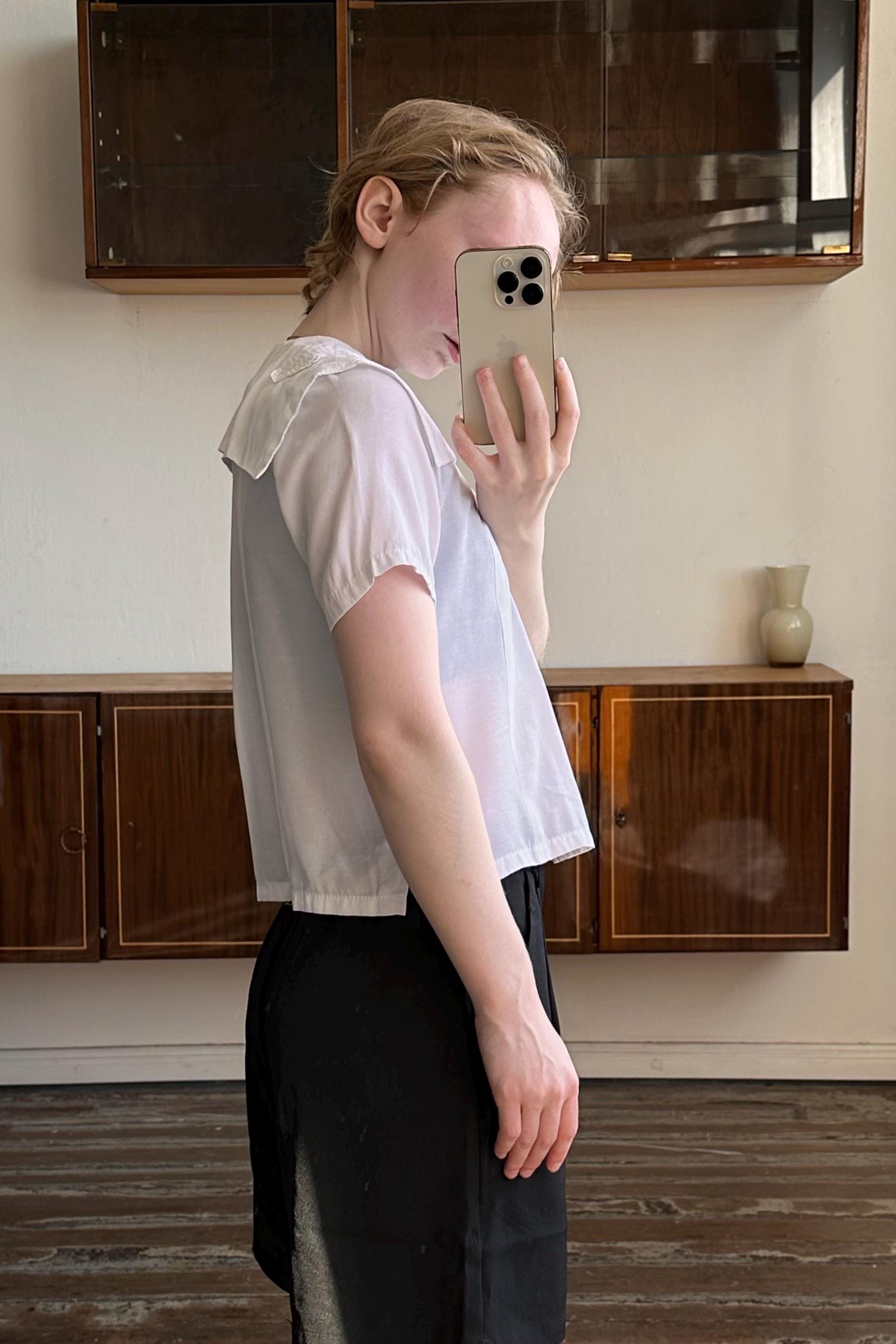 A person is taking a selfie with a white iPhone in a room with wooden cabinets and a vase on a shelf.
