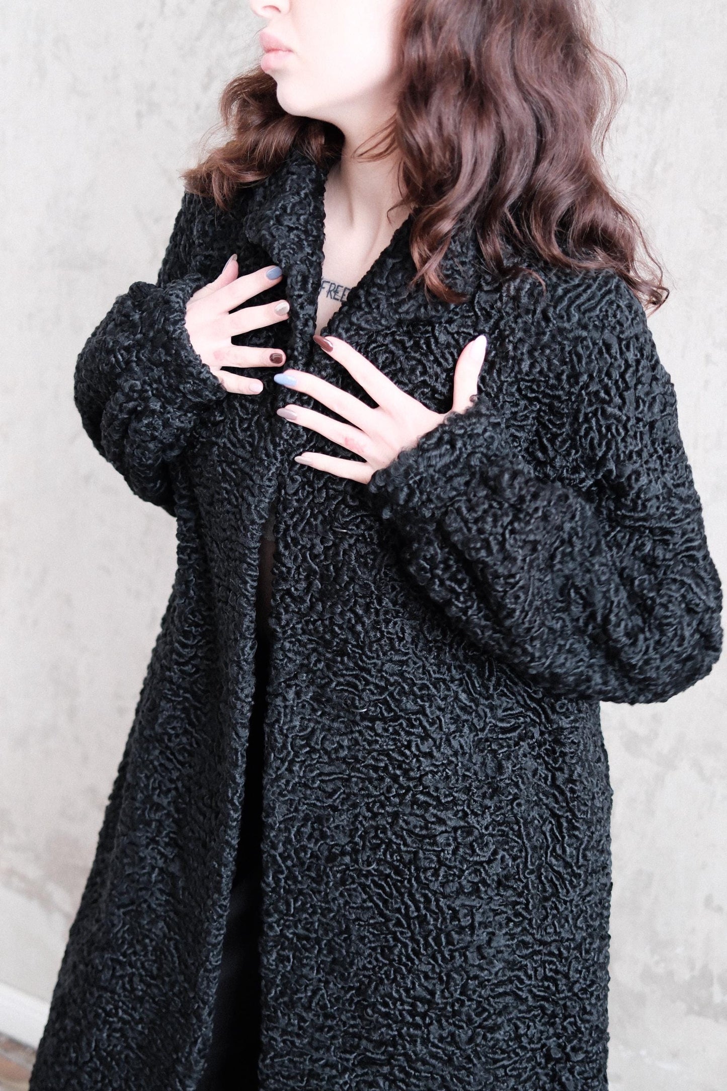 A woman wearing a black, furry coat is lying down on a concrete floor, with her hands raised to her chest.