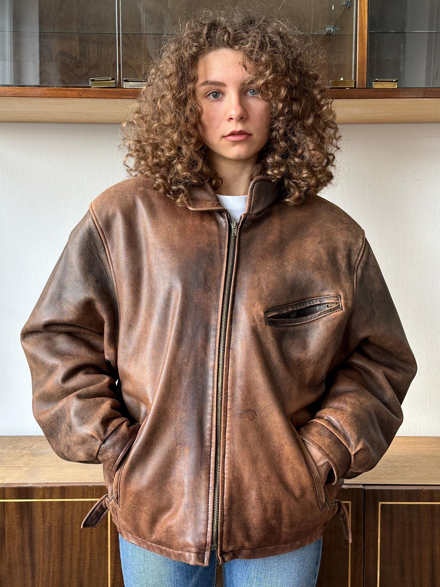 A young woman with curly hair wearing a brown leather jacket stands in front of a wooden cabinet.