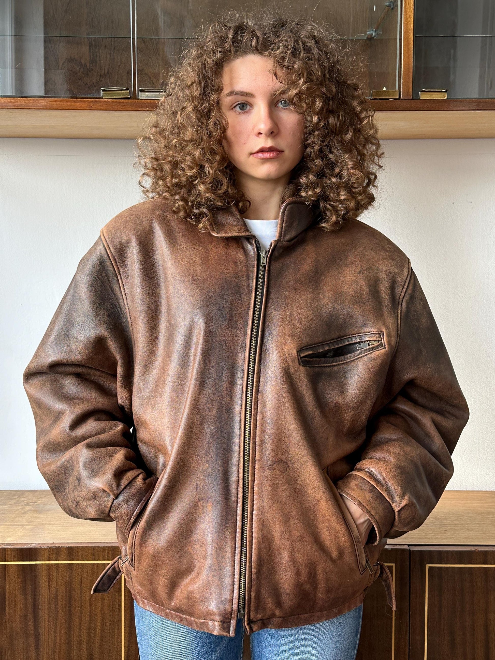 A young woman with curly hair wearing a brown leather jacket stands in front of a wooden cabinet.