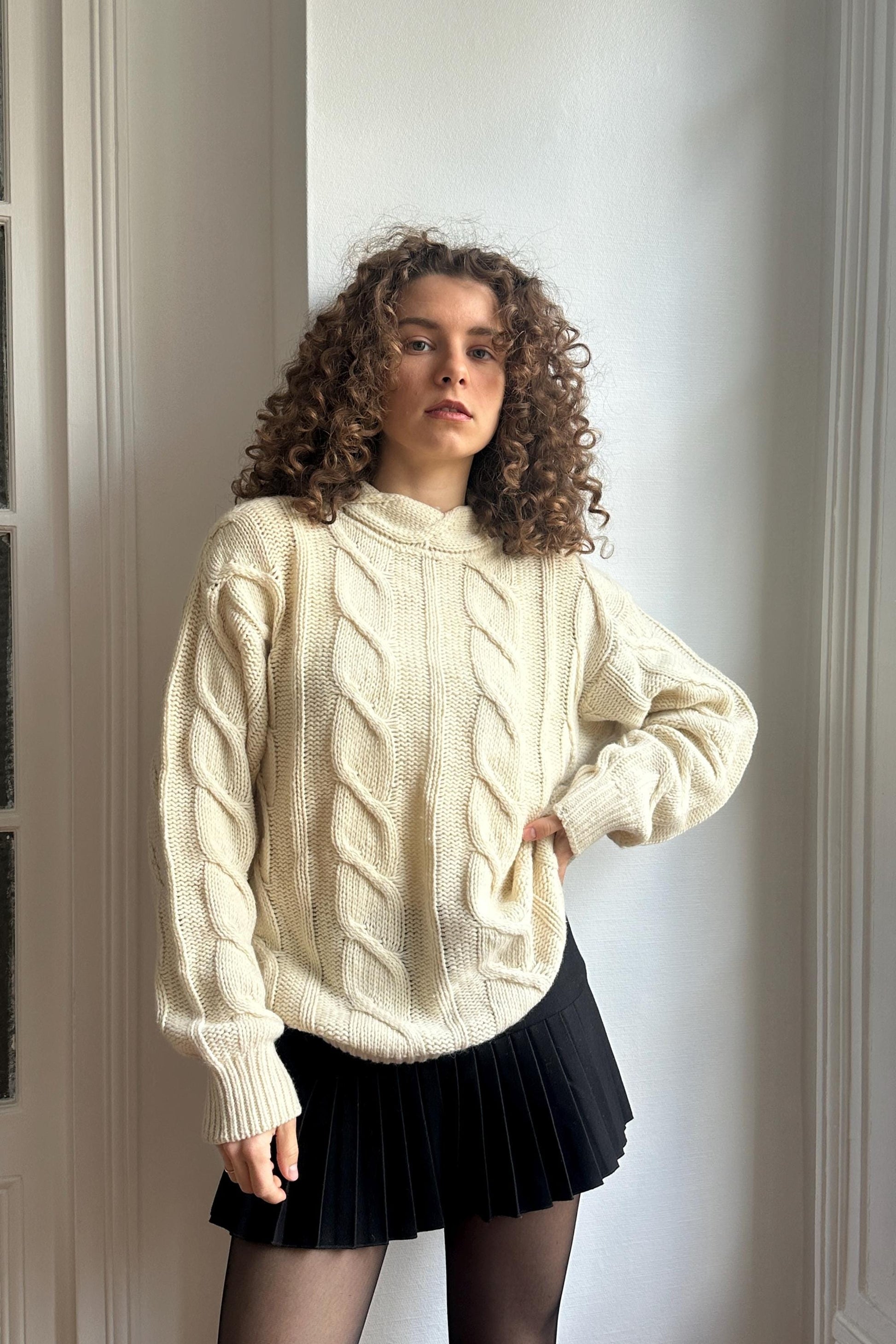 A young woman with curly hair stands confidently in front of a white wall, wearing a cream-colored sweater and a black pleated skirt.
