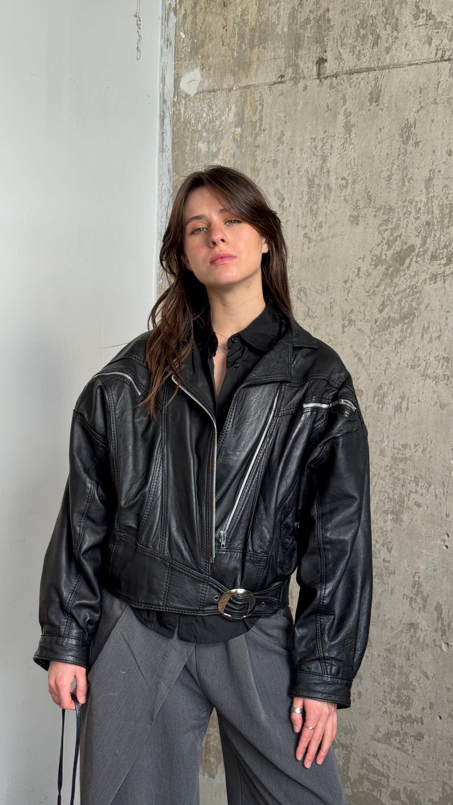 A woman wearing a black leather jacket stands in front of a concrete wall, looking directly at the camera with a neutral expression.