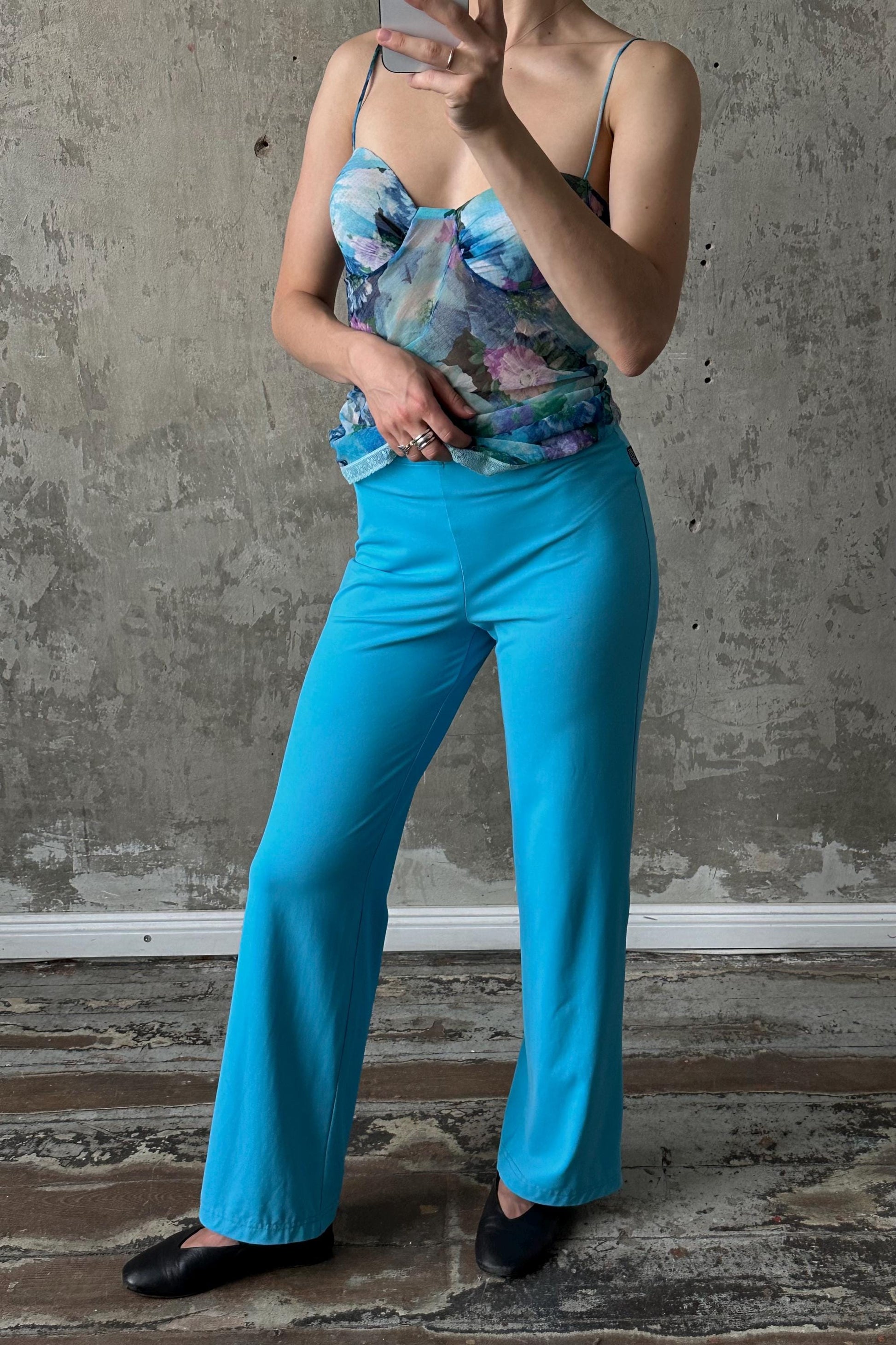 A woman wearing a blue floral top and wide-leg pants is taking a selfie with her phone.