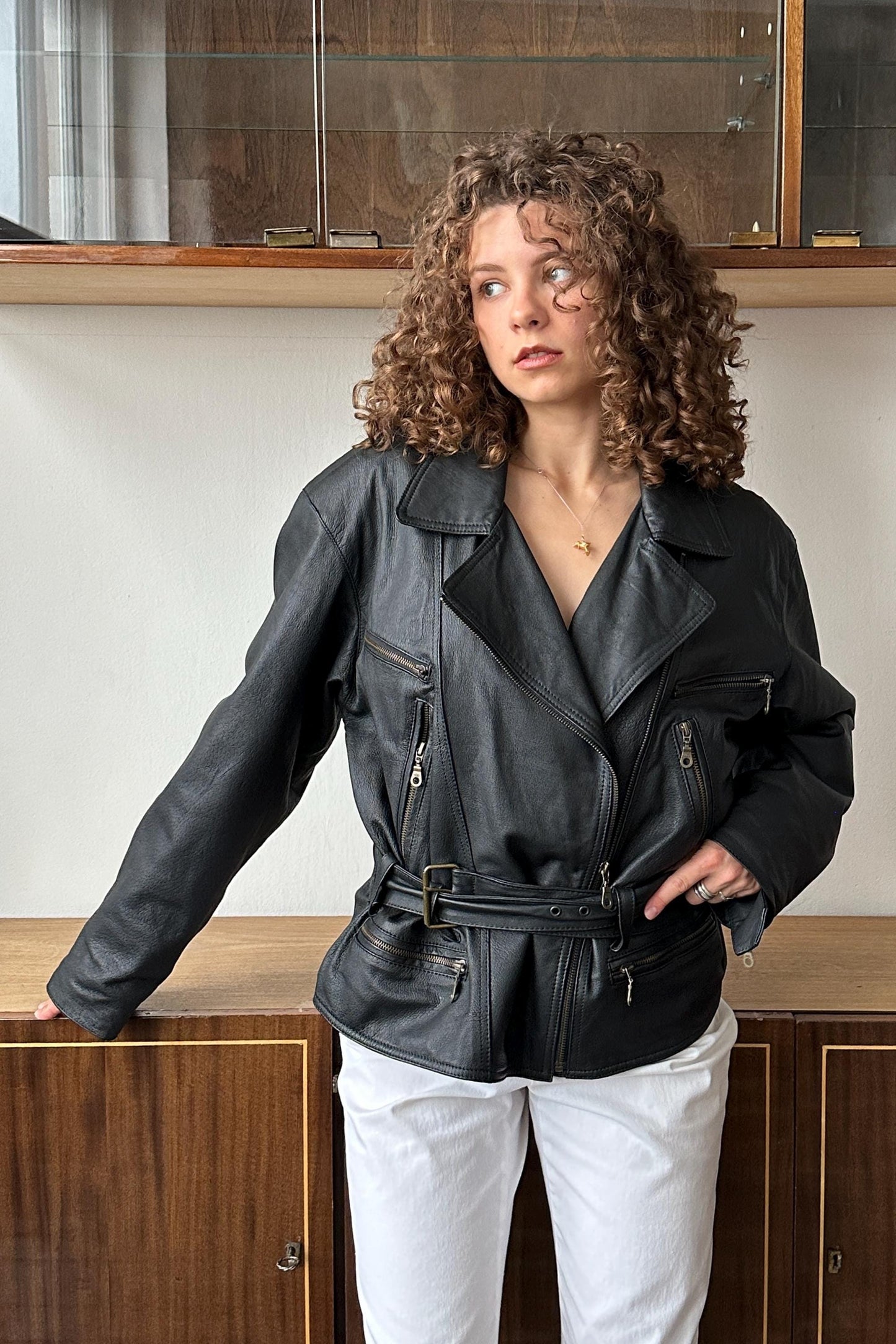 A woman with curly hair wearing a black leather jacket stands in front of a wooden cabinet.