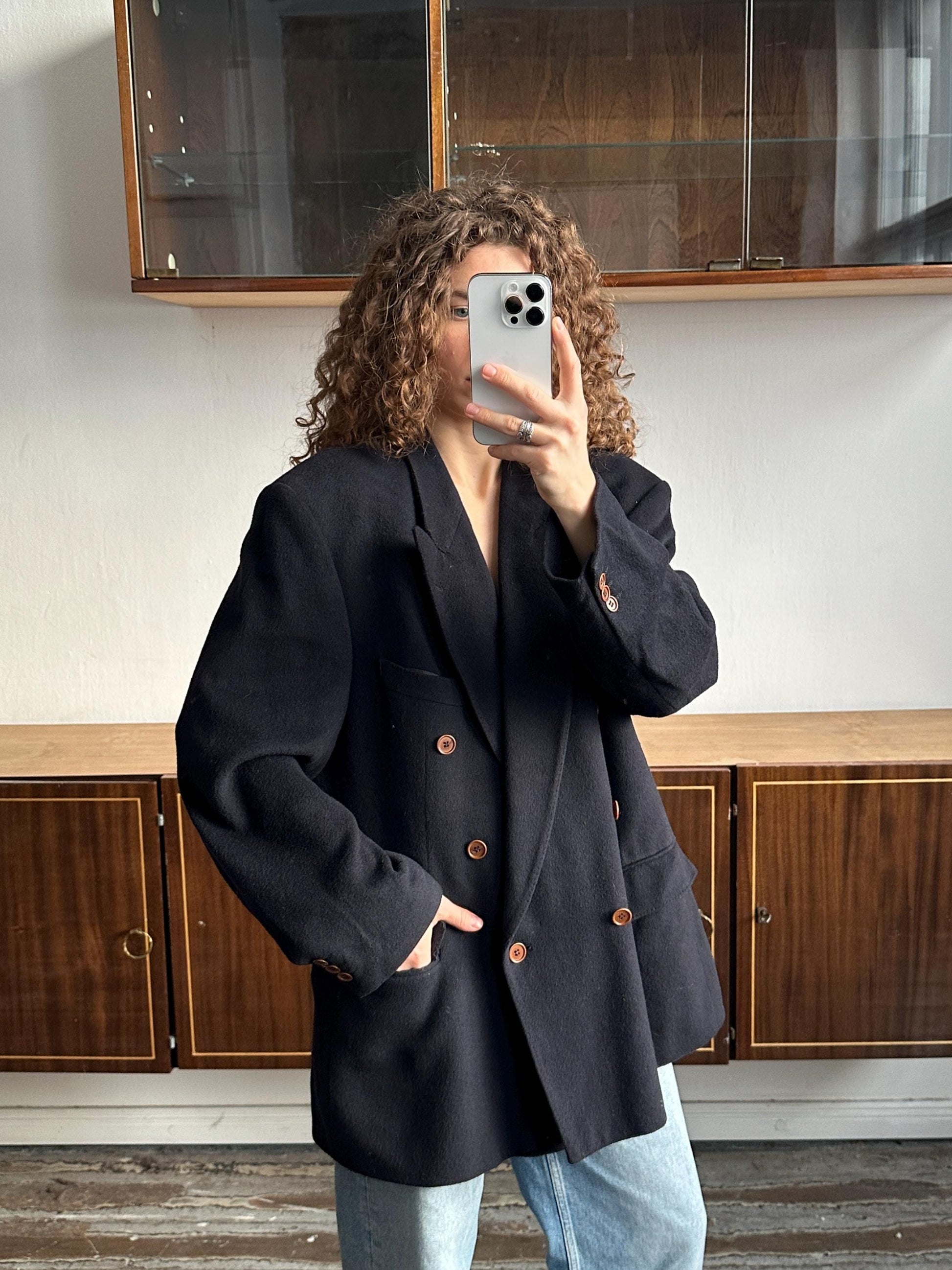 A person with curly hair is taking a selfie with a smartphone, wearing a black blazer.