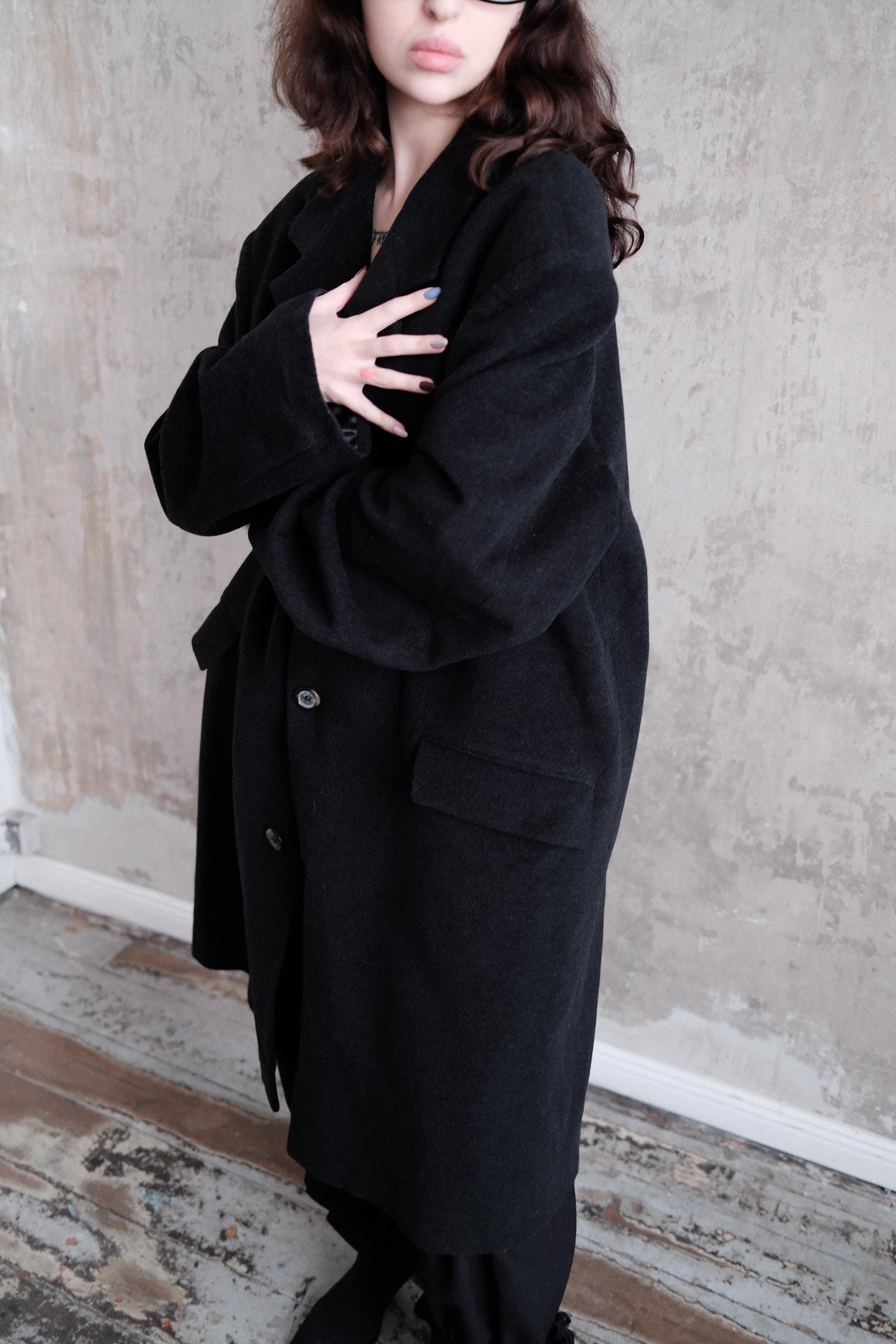 A person wearing a black coat stands against a concrete wall.