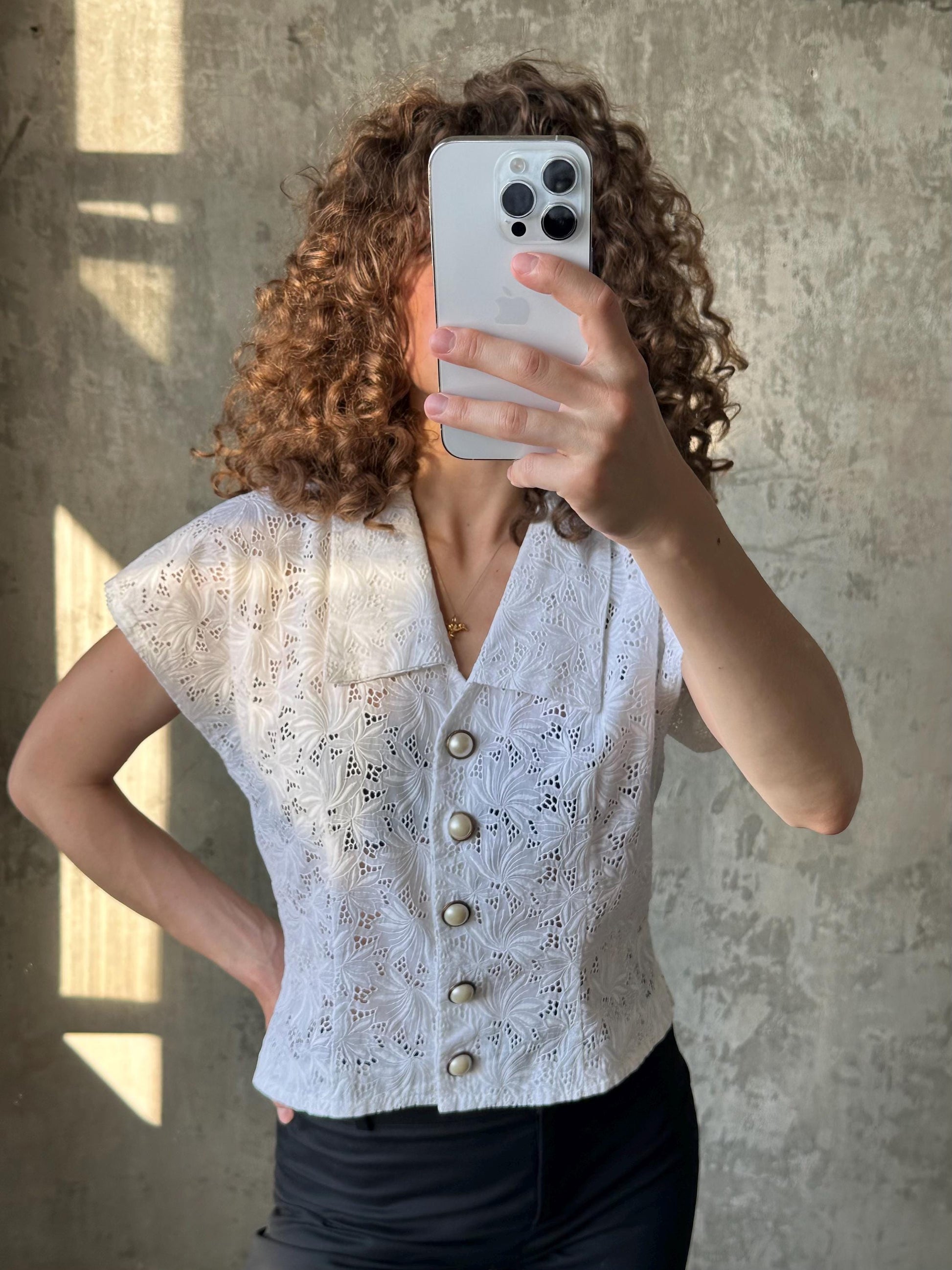 A woman with curly hair is taking a selfie with a white iPhone, wearing a white lace blouse with buttons.