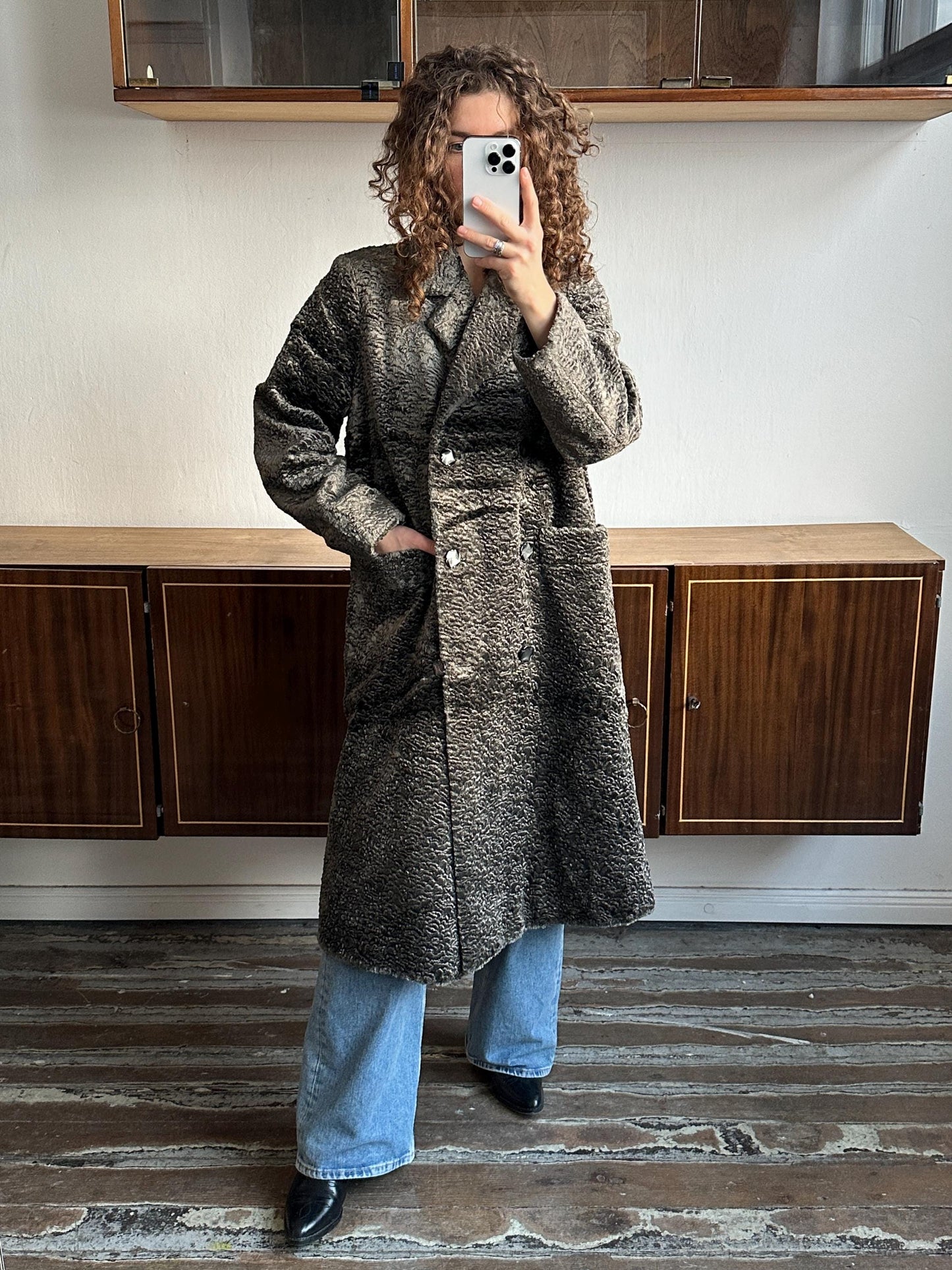 A person is taking a selfie with a smartphone, wearing a long gray coat and blue jeans.