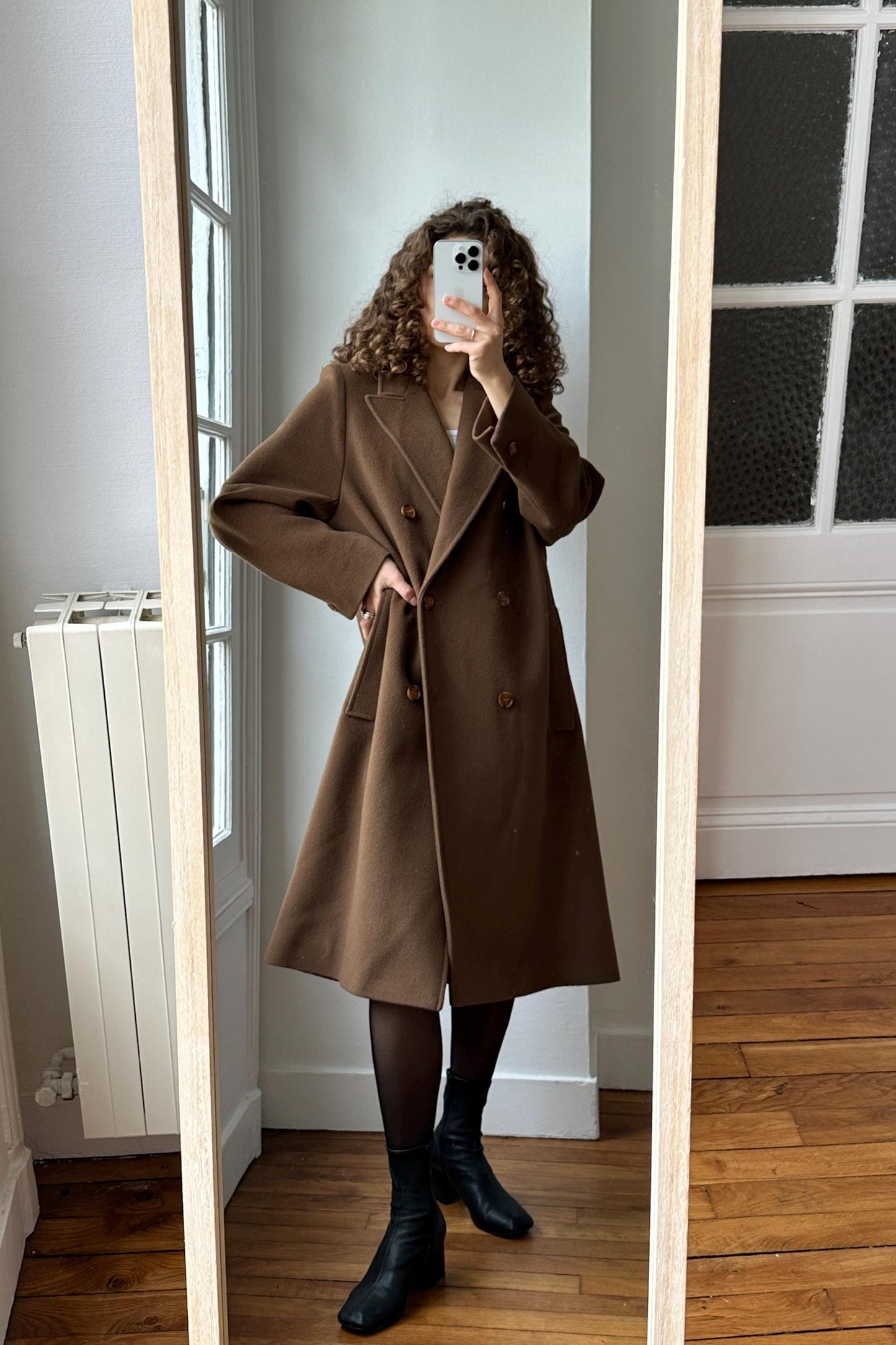 A woman wearing a brown coat is taking a selfie in a mirror.