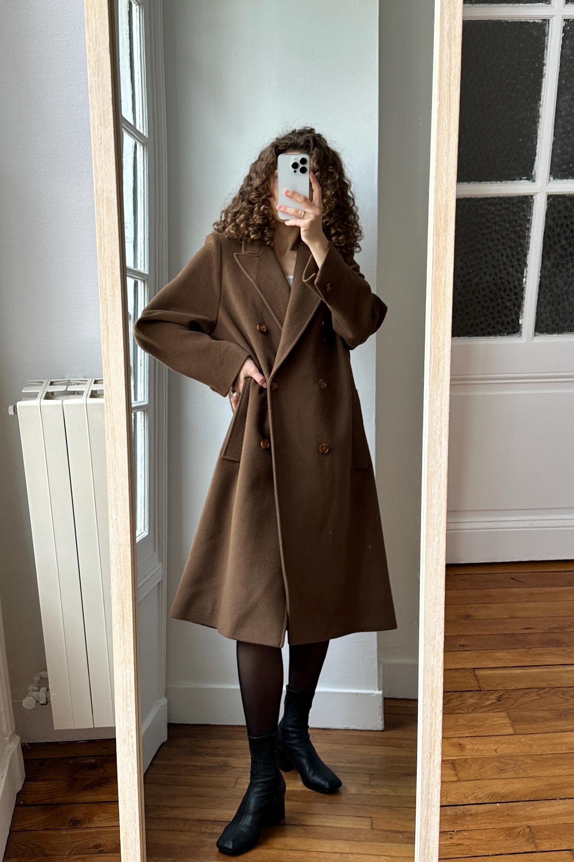 A woman wearing a brown coat is taking a selfie in a mirror.