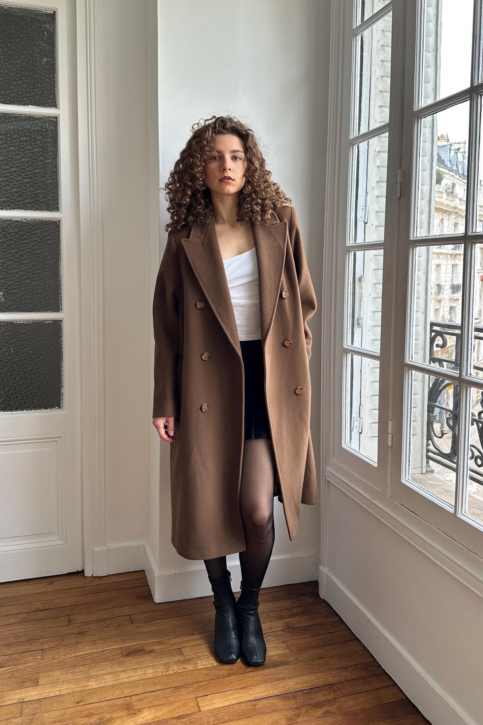 A woman with curly hair stands in a room, wearing a brown coat and black boots.